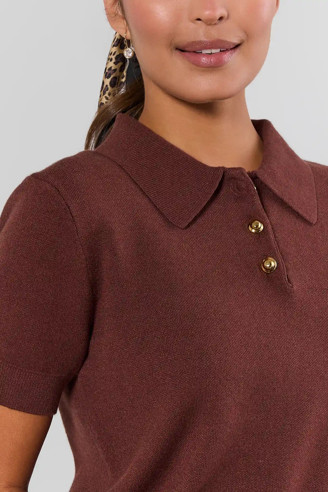 Close-up of a woman wearing a short-sleeve knit polo shirt in a warm rust brown color, featuring a classic collar and two gold-tone buttons.
