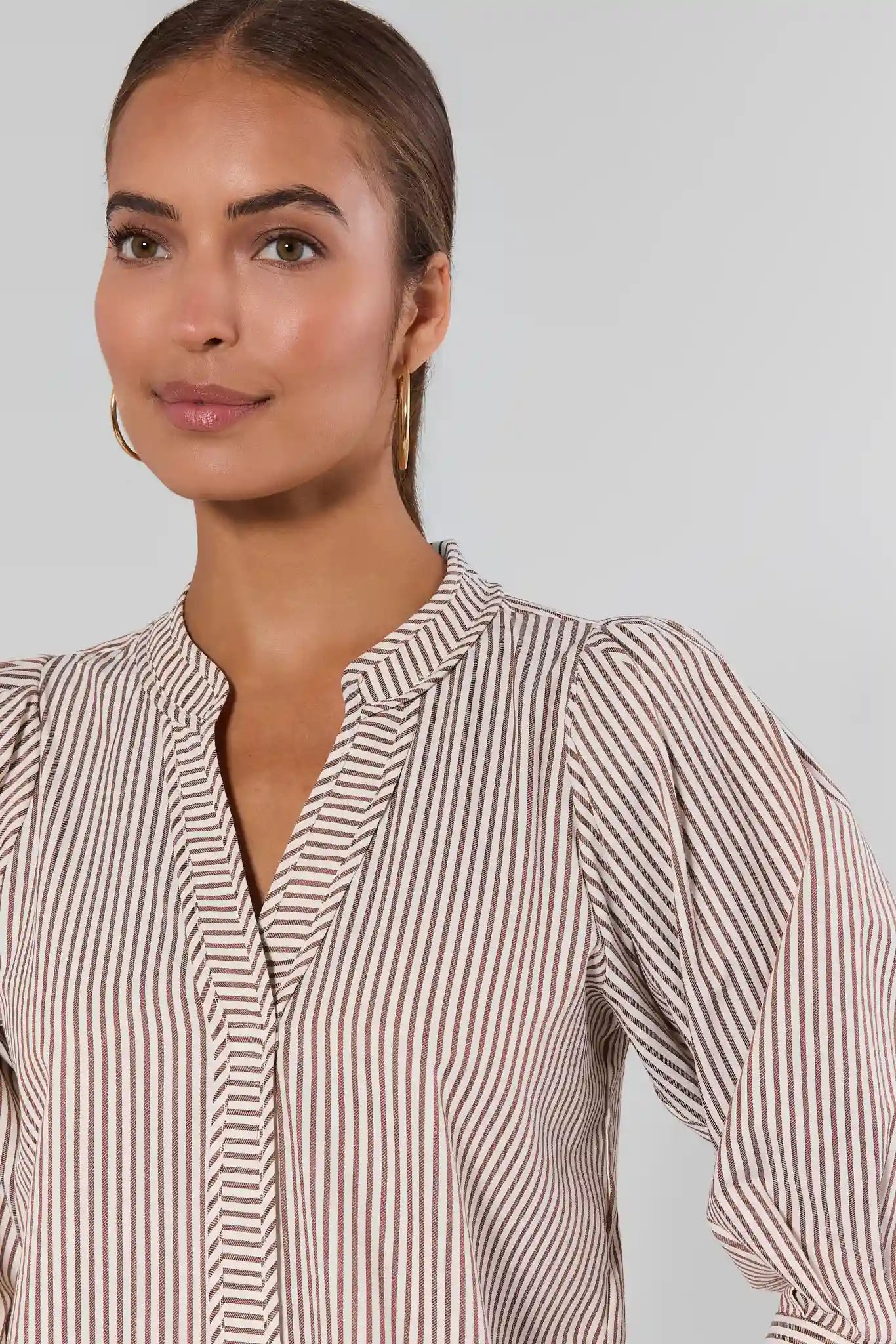 A woman wearing a beige and brown vertically striped long-sleeve blouse with a split neckline, shown against a plain light gray studio background.