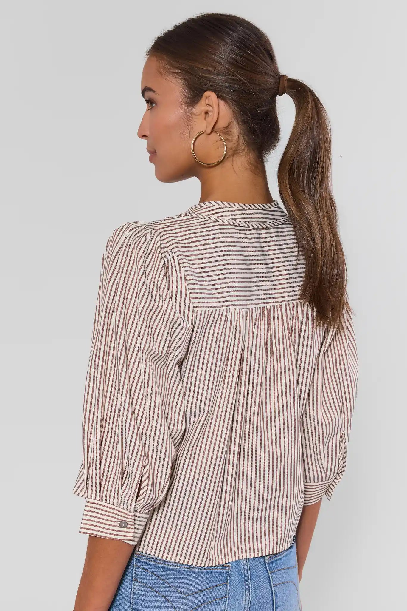 A woman shown from the back wearing a brown and white vertically striped blouse with puffed three-quarter sleeves and light blue jeans against a light gray studio background.