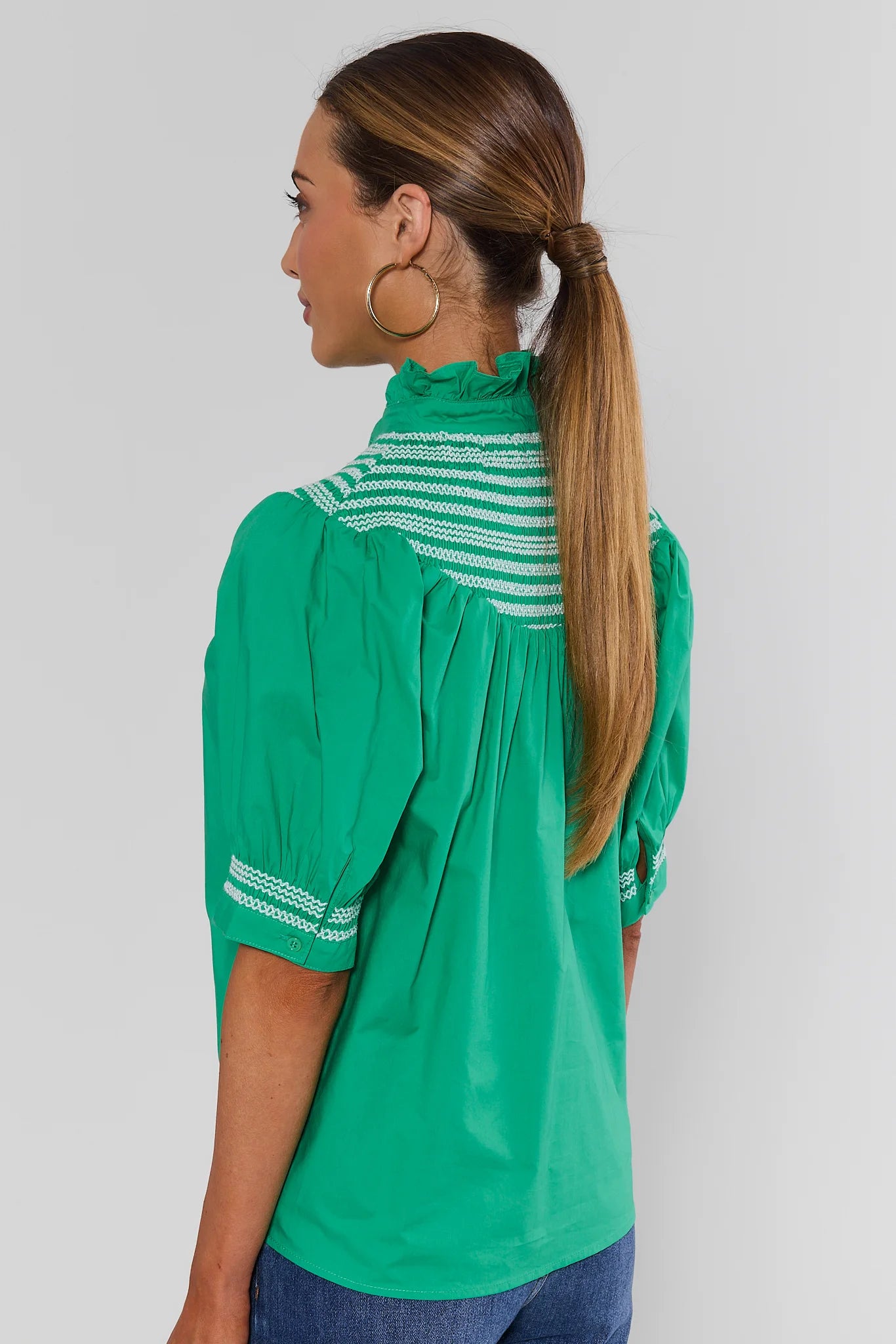 A woman shown from the back wearing a bright green blouse with white striped yoke detailing and puff sleeves, paired with blue jeans against a light gray studio background.