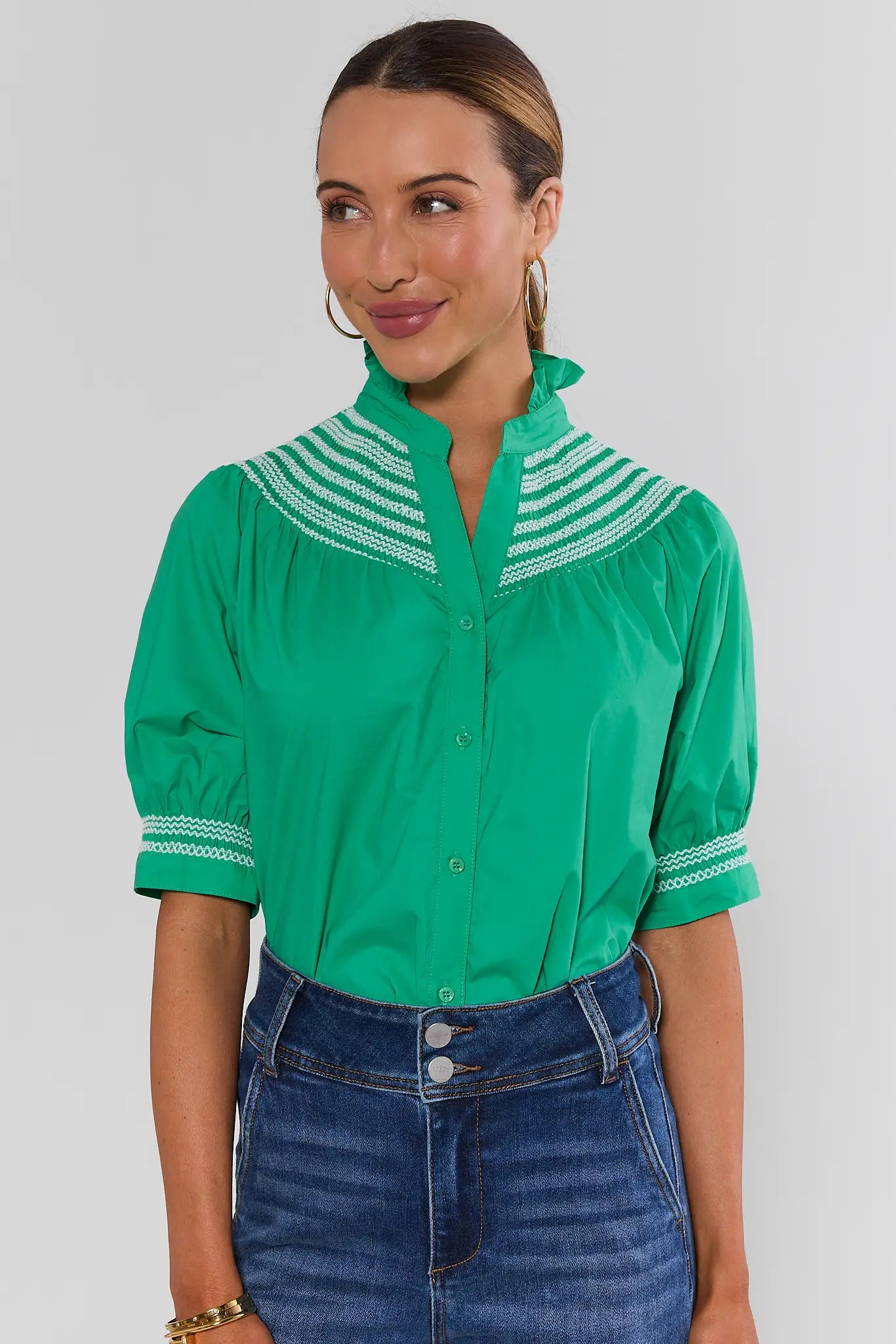 A woman wearing a bright green short-sleeve button-up blouse with white striped embroidered details on the collar and cuffs, paired with a blue denim skirt, against a light gray studio background.
