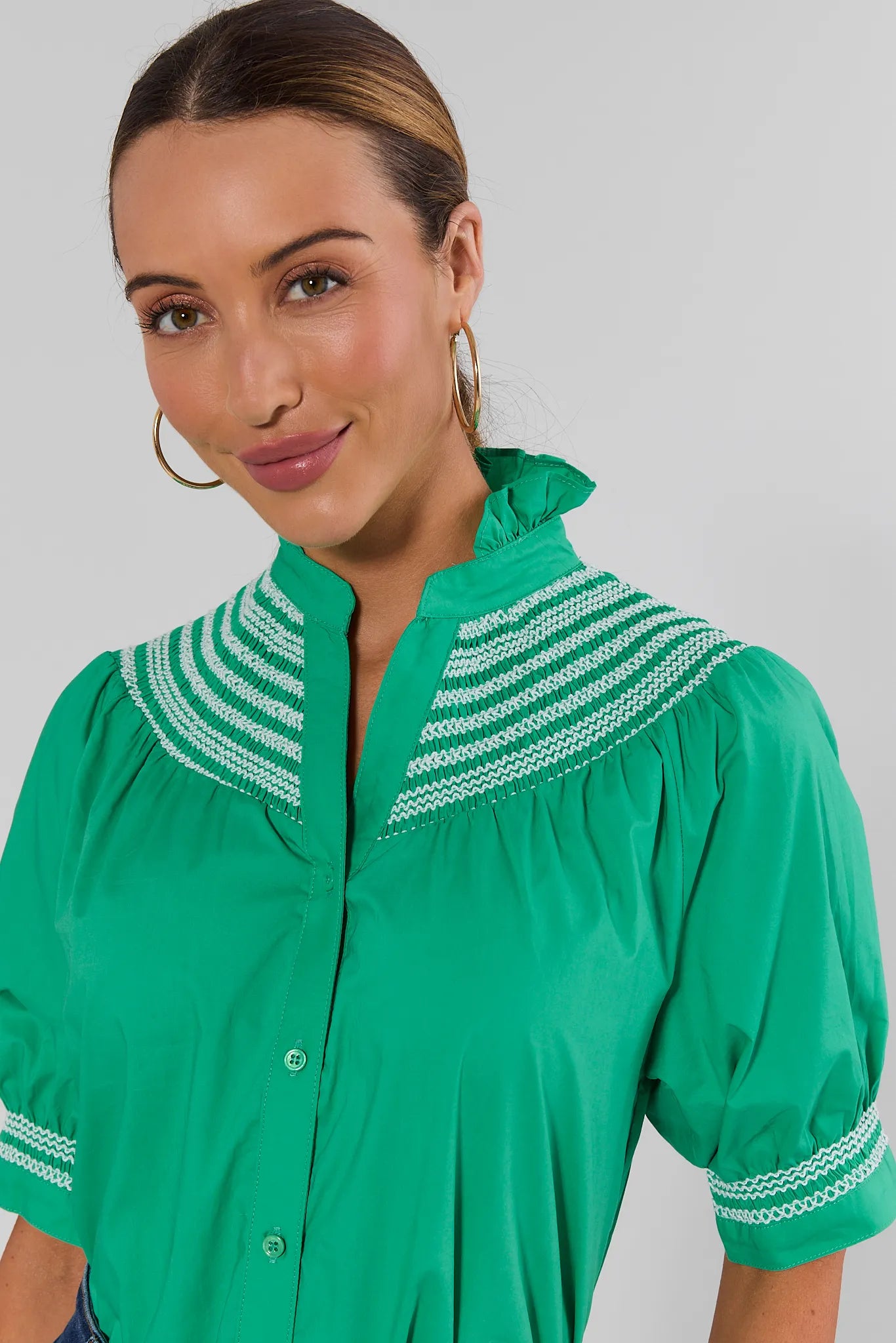 A woman wearing a bright green short-sleeve blouse with white embroidered stripes around the collar and cuffs, photographed against a light gray studio background.