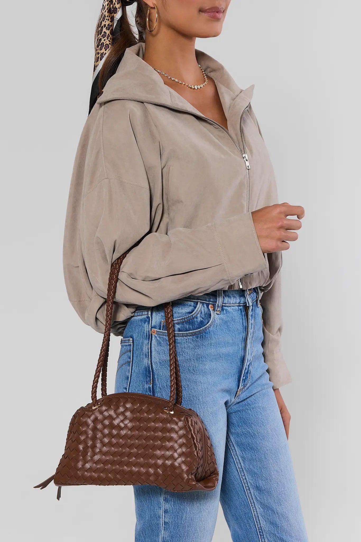 A woman wearing a taupe zip-up hooded jacket and blue jeans, carrying a brown woven leather dome handbag with braided shoulder straps against a light gray studio background.
