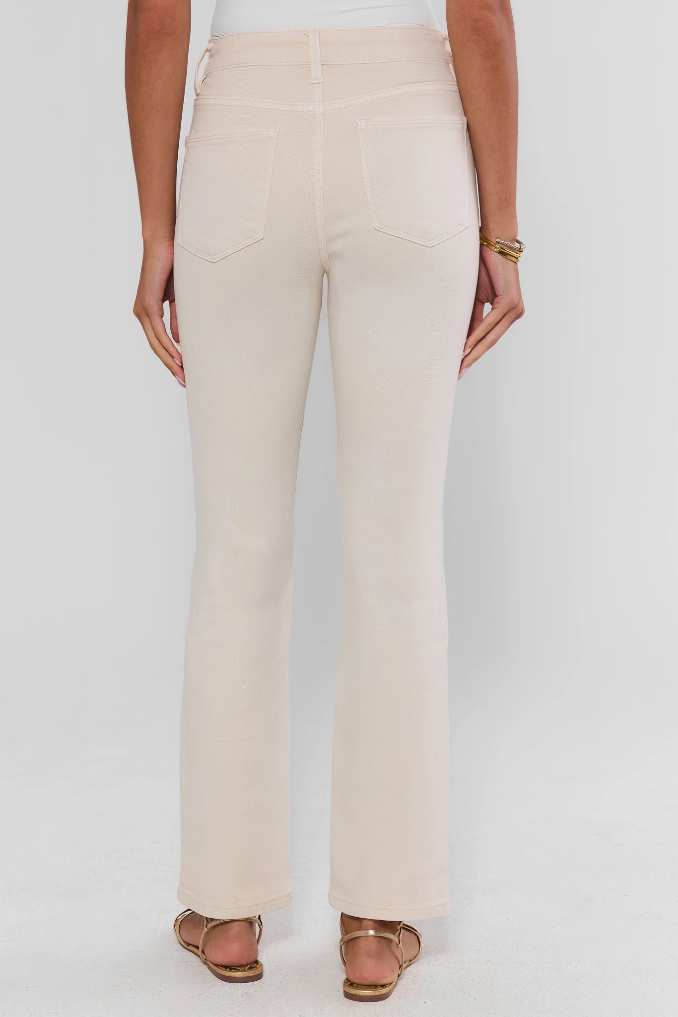 A person shown from the back wearing light beige straight-leg jeans with a white top and brown sandals, against a plain light gray studio background.