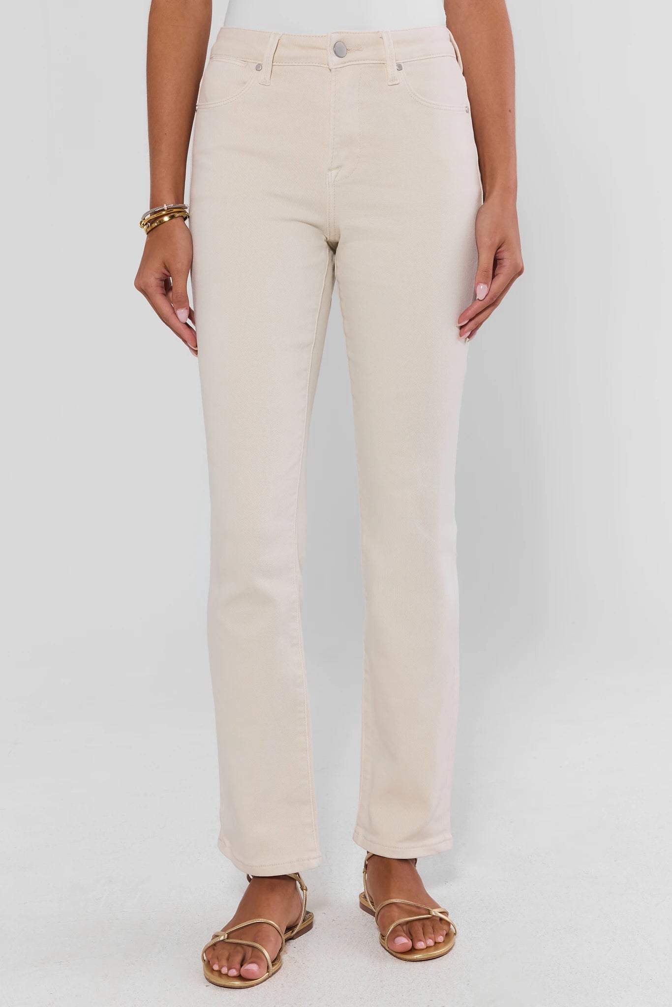 A model is wearing a pair of straight-leg, high-rise cream-colored denim jeans with a button fly and belt loops, paired with flat brown sandals against a light gray studio background.