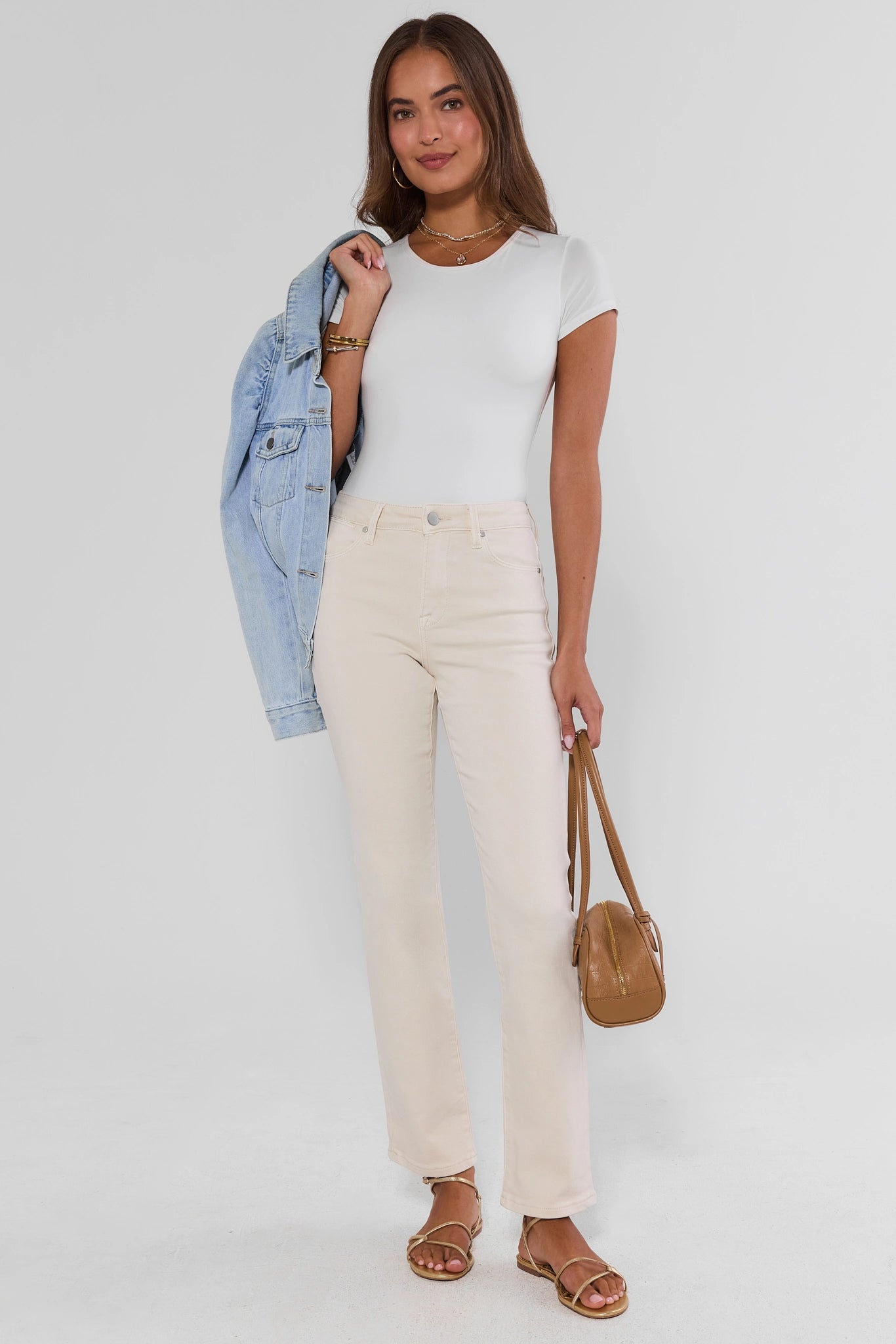 A woman modeling a white short-sleeve crewneck t-shirt, cream-colored straight-leg jeans, a light blue denim jacket draped over her arm, and brown leather sandals, holding a small brown leather handbag against a plain light gray studio background.