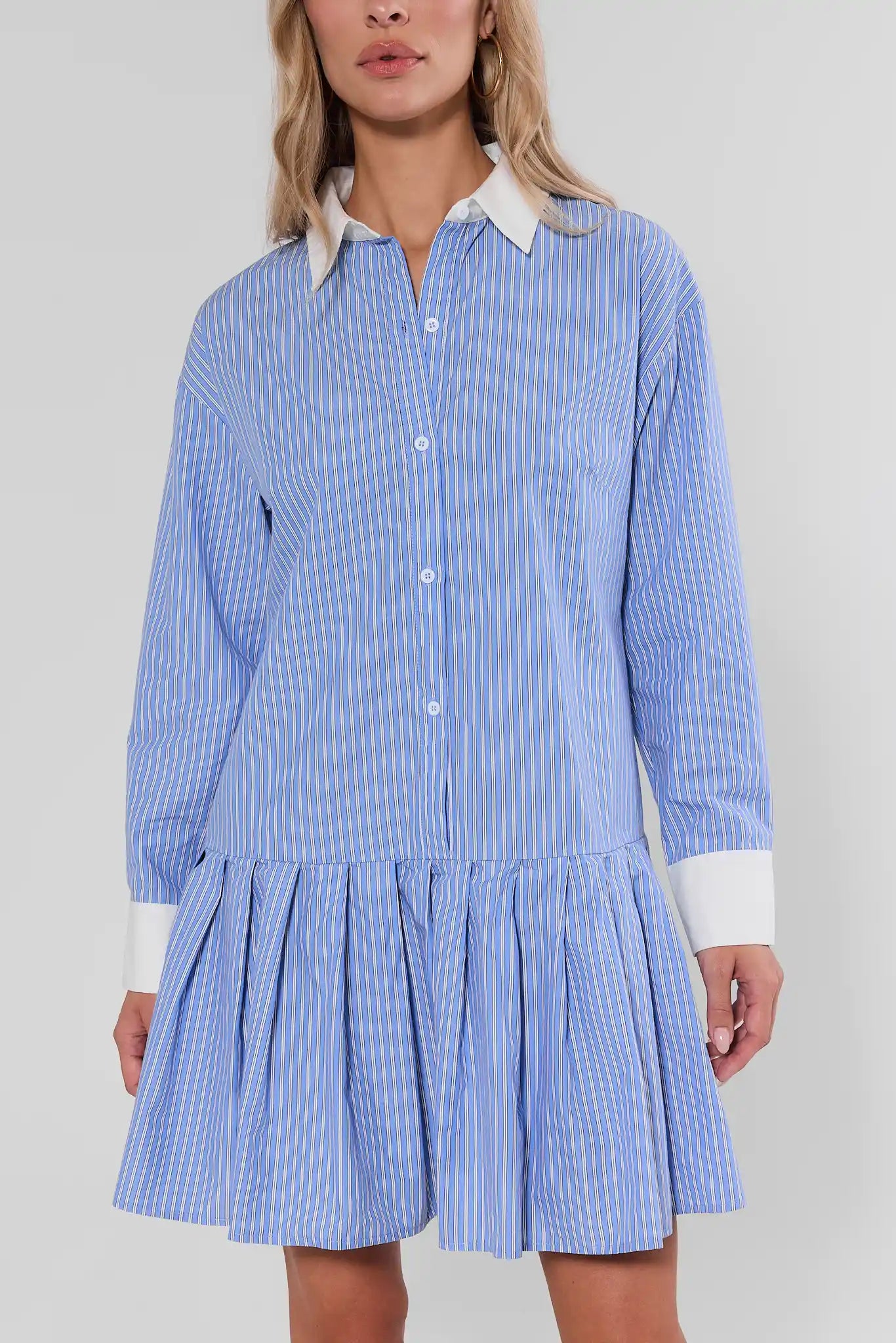 A woman wearing a light blue and white vertically striped shirt dress with a white pointed collar and white cuffs, shown against a plain light gray studio background.