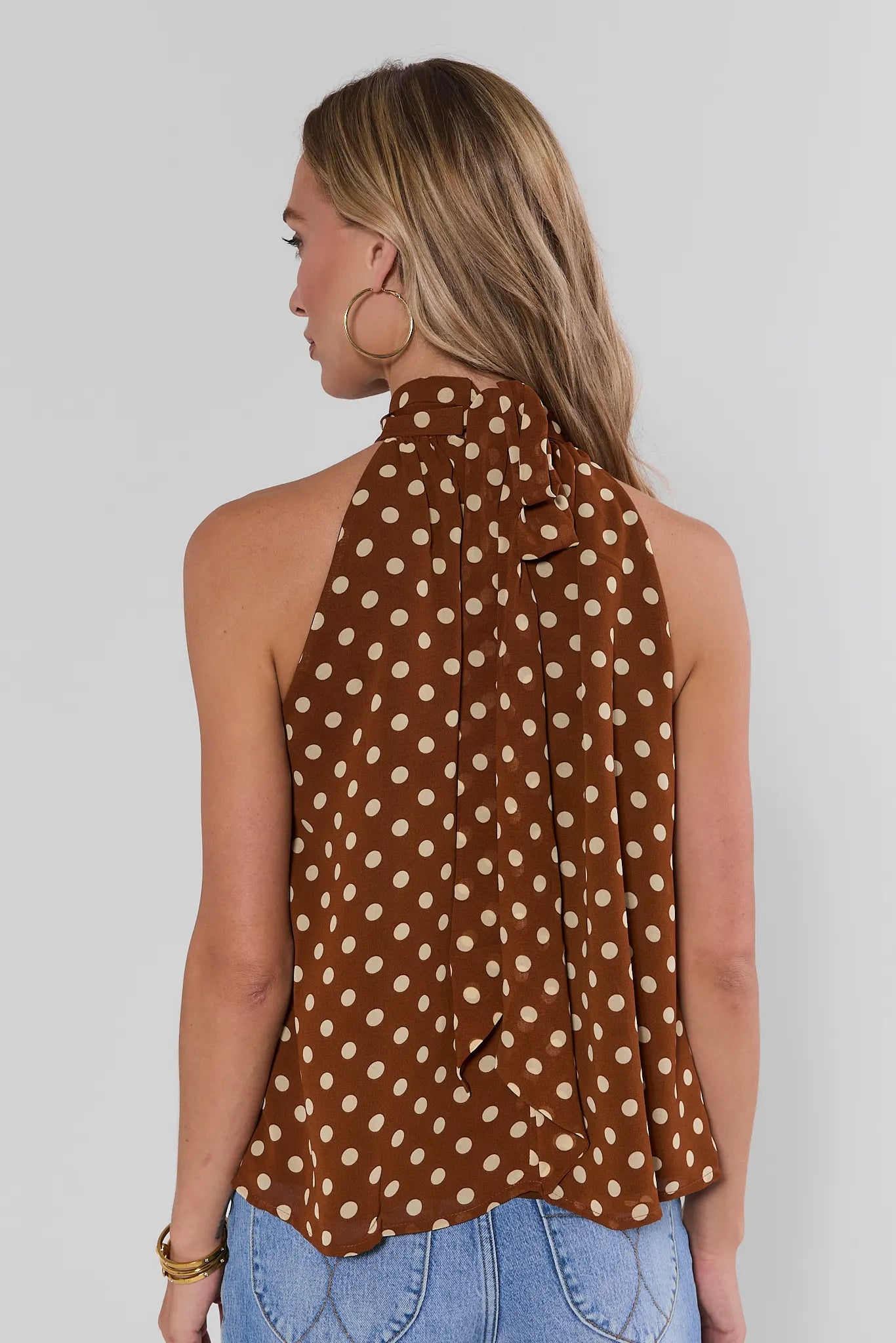 A woman shown from the back wearing a brown halter-style top with a beige polka dot pattern, paired with light blue jeans, against a plain light gray background.
