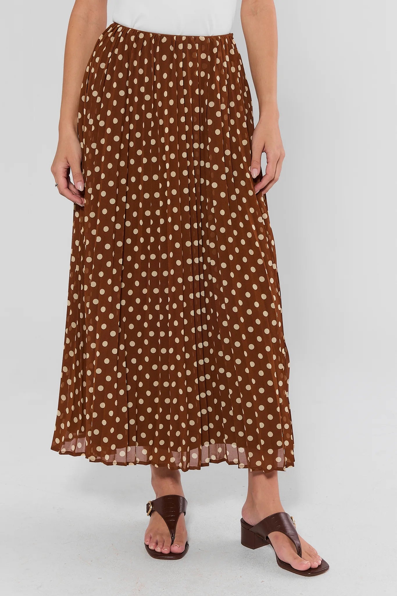 A brown pleated maxi skirt with a beige polka dot pattern, worn by a model with brown sandals against a light gray background.