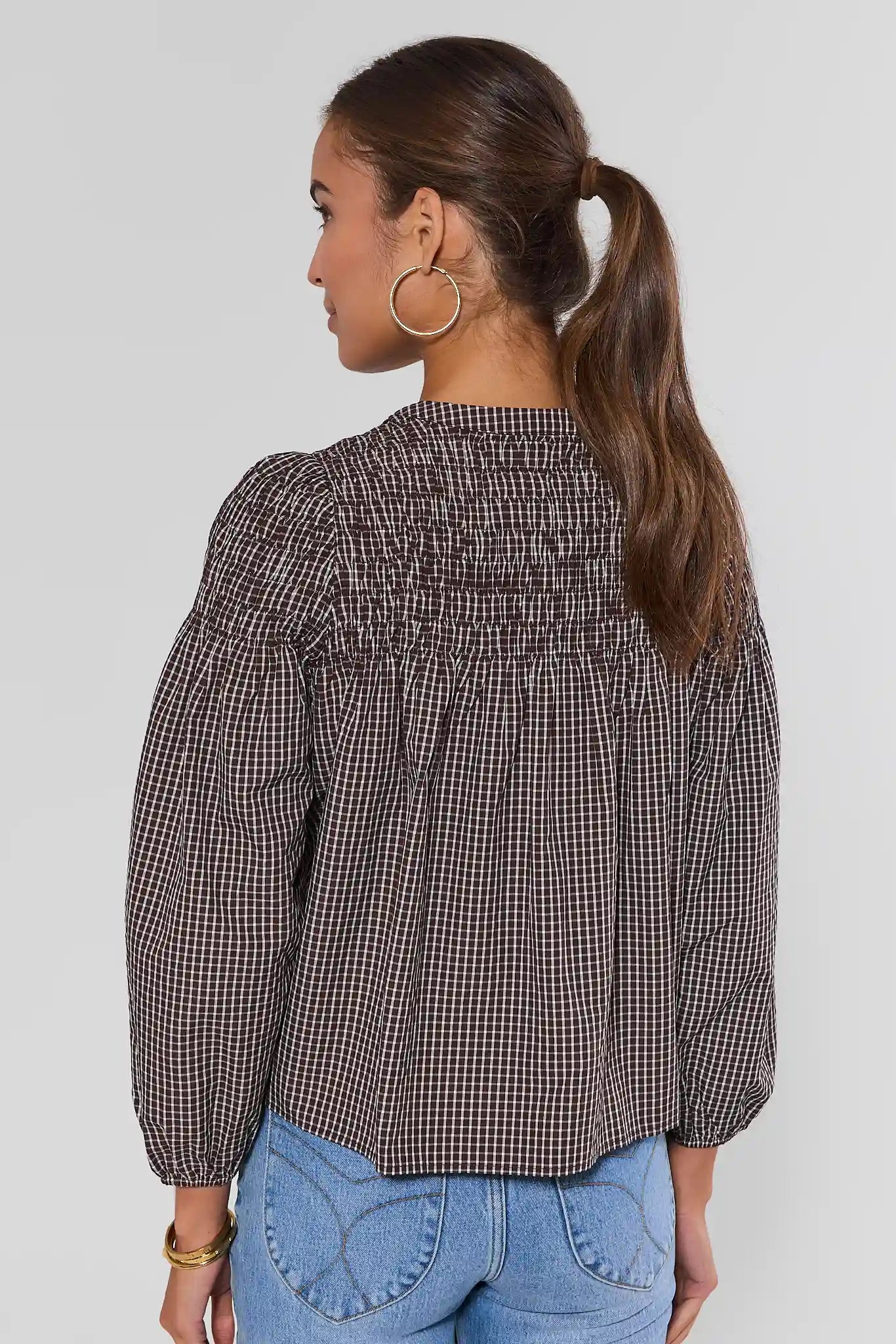 A woman shown from the back wearing a brown and black gingham long-sleeve blouse with a smocked bodice and puffed sleeves, paired with light blue jeans against a light gray studio background.