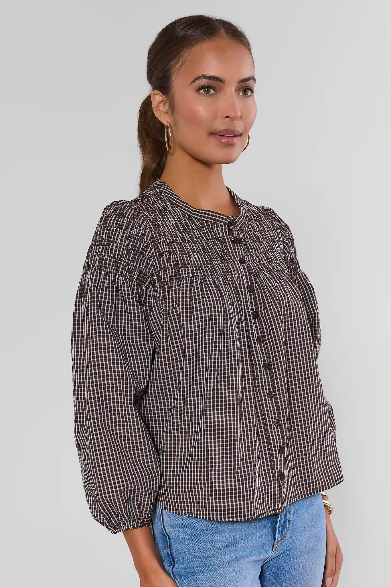 A woman wearing a brown and white checkered long-sleeve blouse with puff sleeves and a button-up front, paired with blue jeans, against a light gray background.