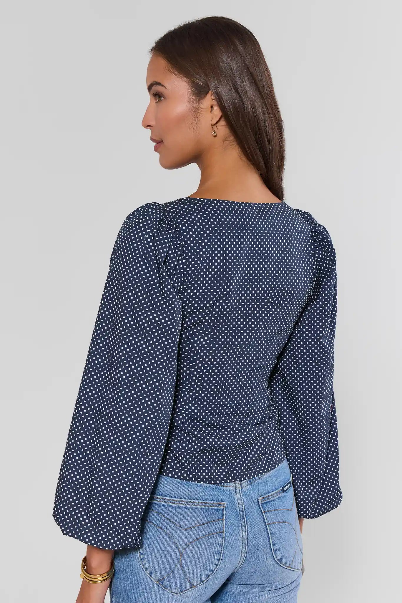 A woman shown from the back wearing a navy blue long-sleeve top with a white polka dot pattern and light blue jeans, against a plain light gray background.