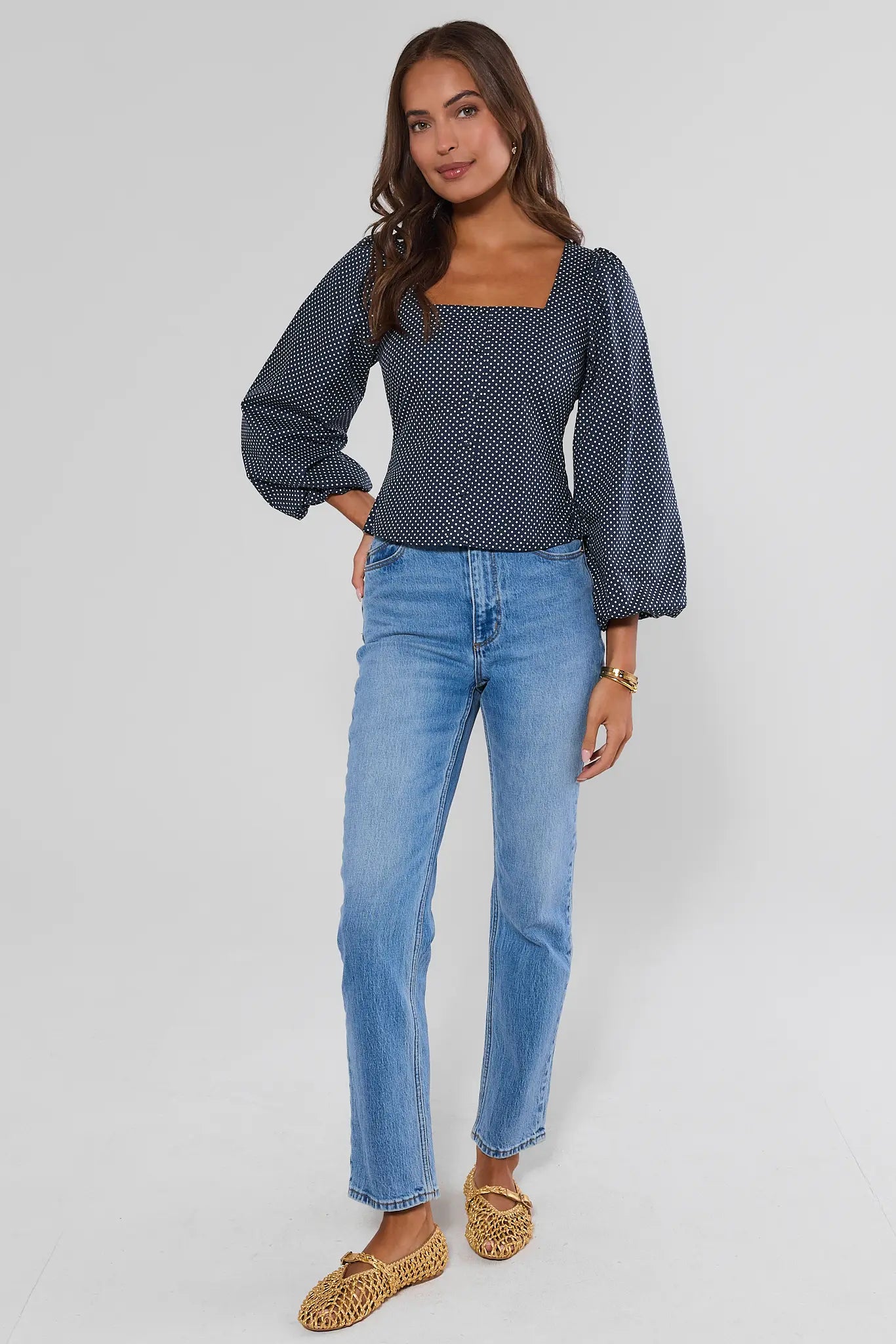 A woman modeling a navy blue and white micro-dot patterned blouse with puff sleeves and a square neckline, paired with light-wash blue jeans and leopard-print flat shoes, against a light gray studio background.