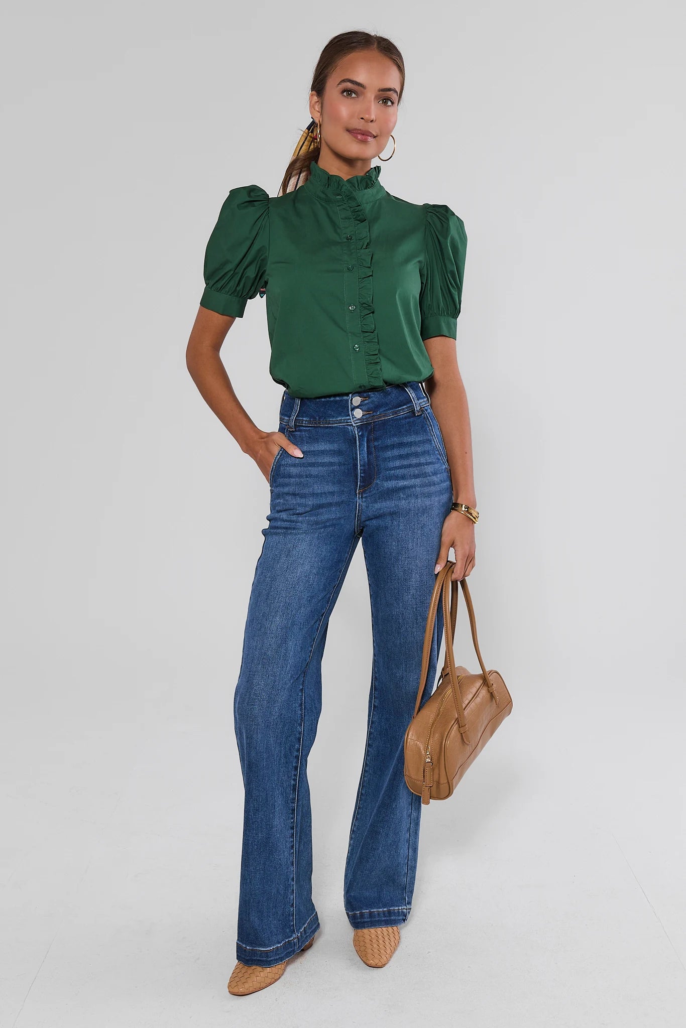A woman modeling a dark green satin puff-sleeve blouse with a high ruffled collar, paired with blue flare jeans, beige pointed-toe flats, and a tan leather handbag, against a light gray studio background.