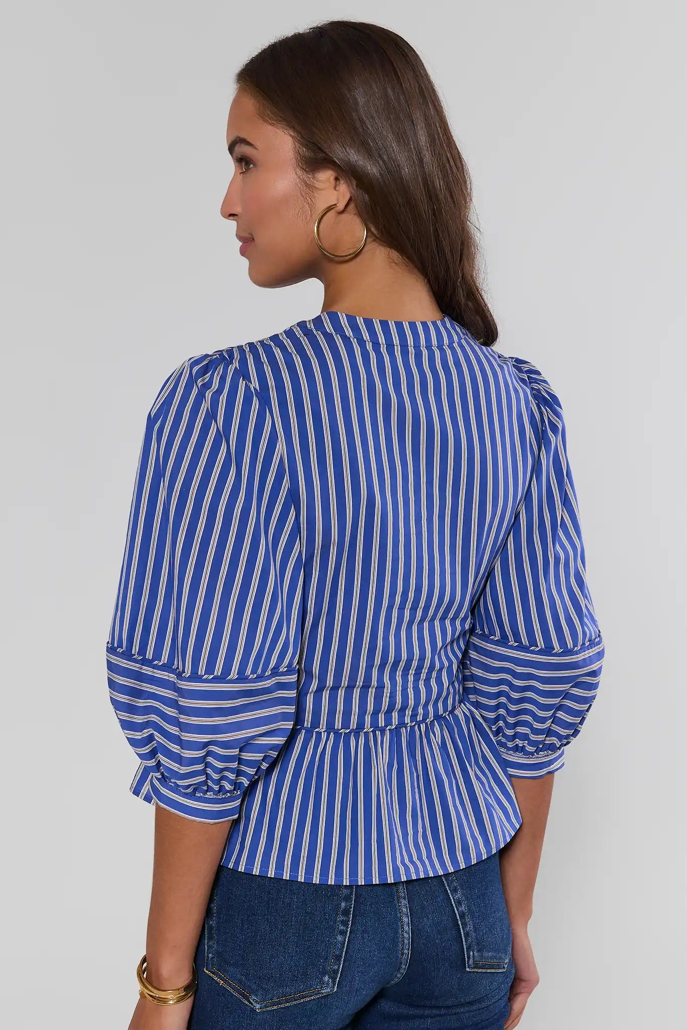 A woman shown from the back wearing a blue and white vertically striped blouse with puffed three-quarter sleeves and a gathered peplum hem, paired with blue jeans against a light gray studio background.