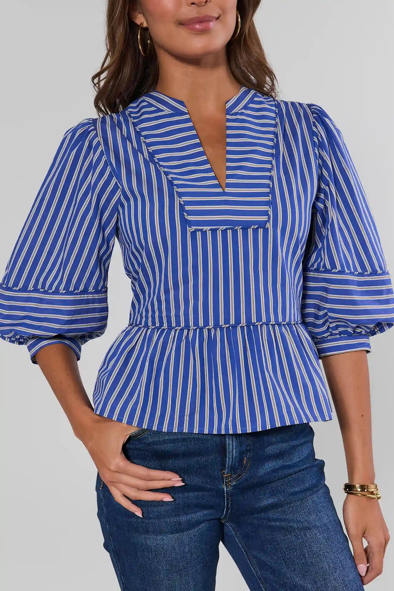 A woman wearing a blue and white vertically striped peplum blouse with puff sleeves and a split V-neckline, paired with blue jeans against a light gray studio background.