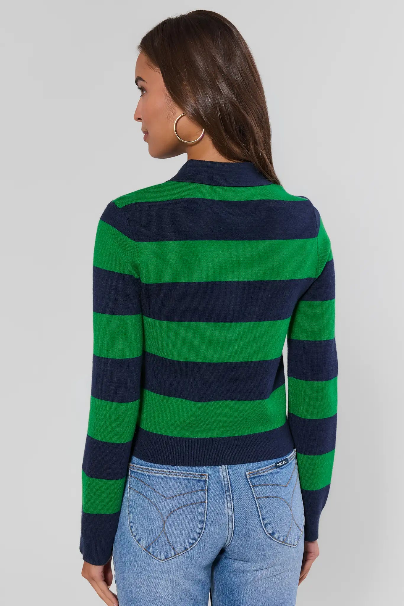 A woman shown from the back wearing a green and navy blue horizontally striped mock neck sweater and light wash blue jeans against a light gray studio background.