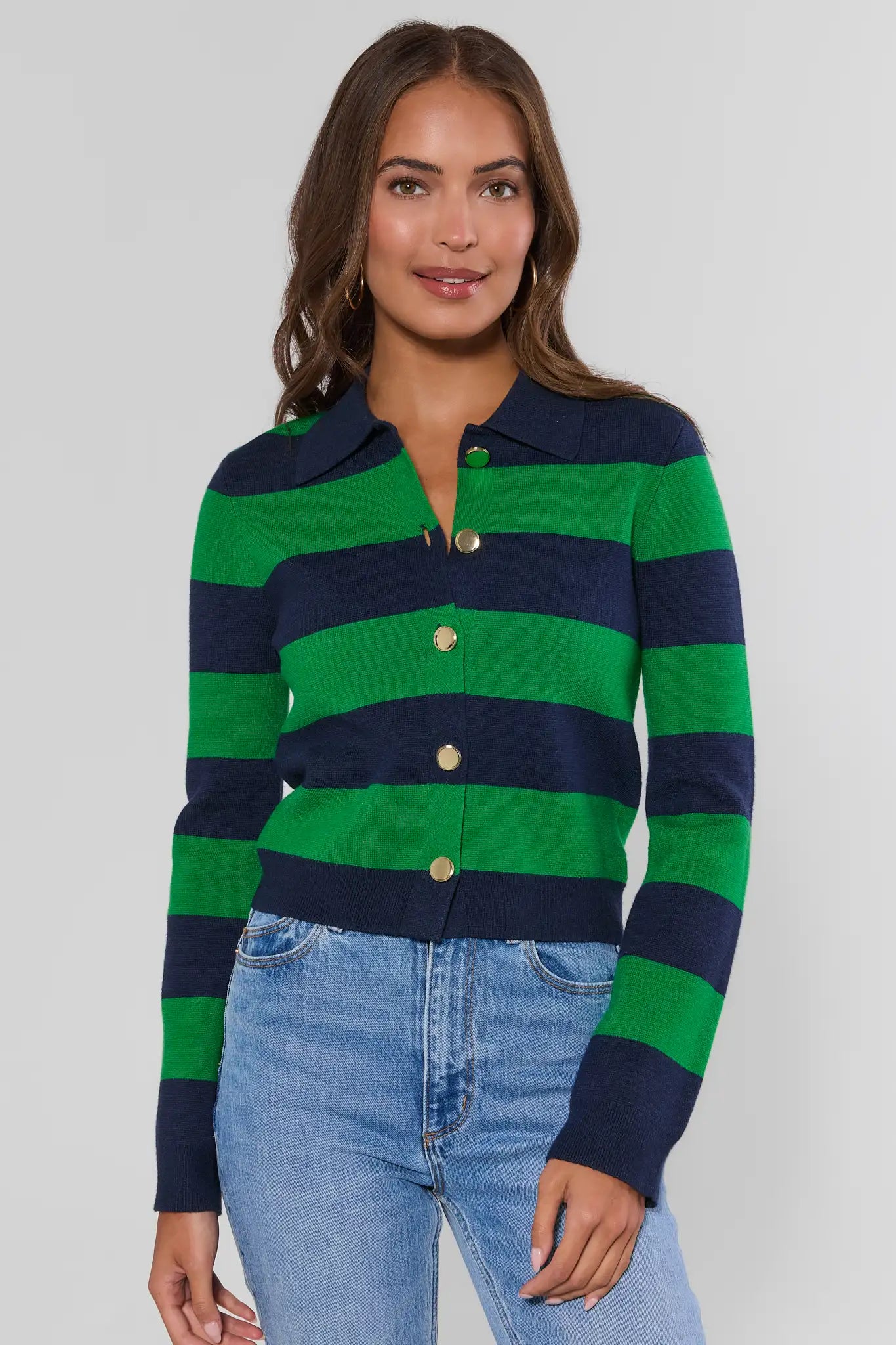 A woman wearing a green and navy blue horizontally striped knit polo cardigan with gold buttons, paired with light wash blue jeans, against a solid light gray studio background.