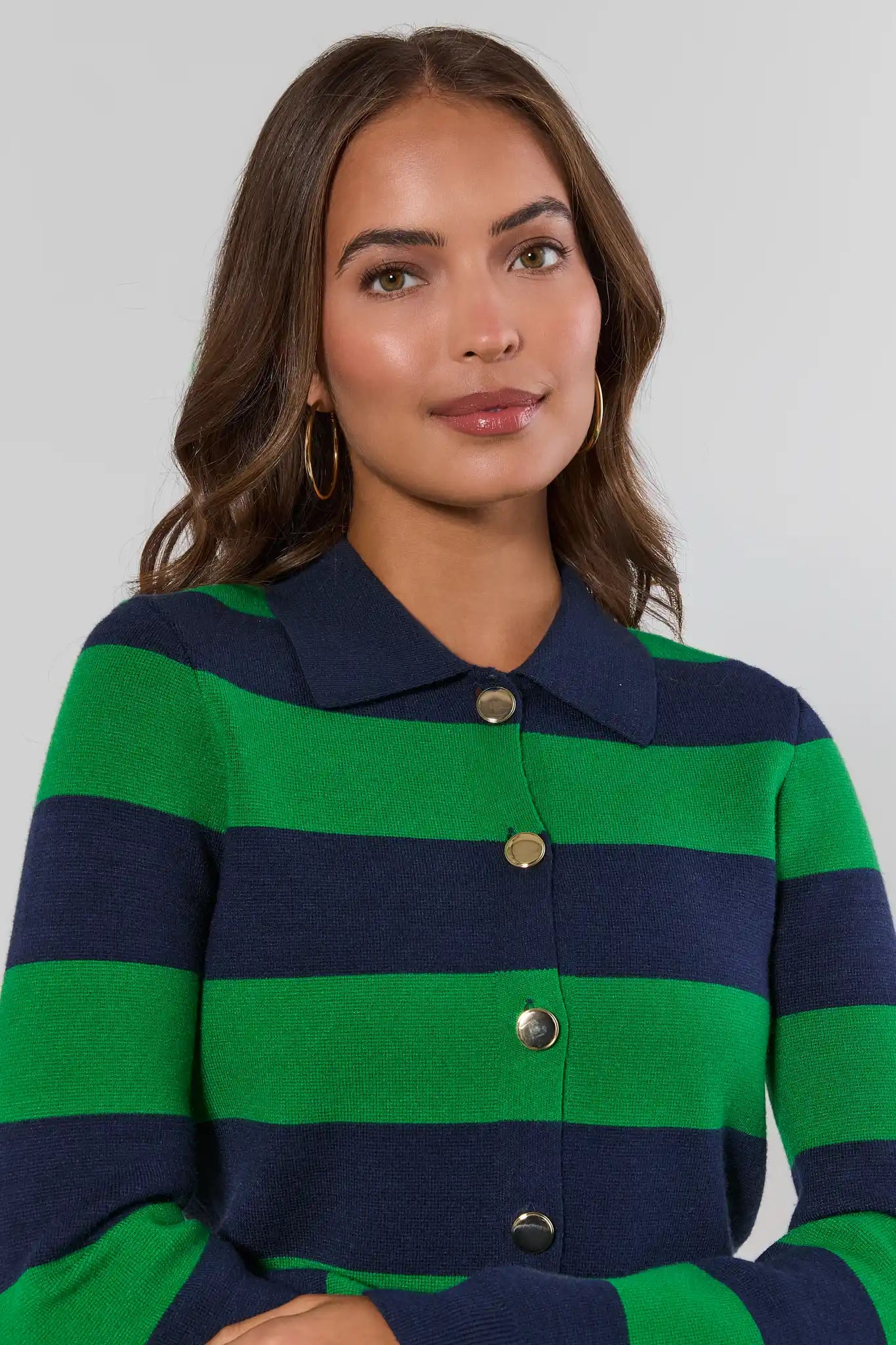 A woman wearing a green and navy blue horizontally striped knit cardigan with a collar and gold buttons, photographed against a light gray studio background.