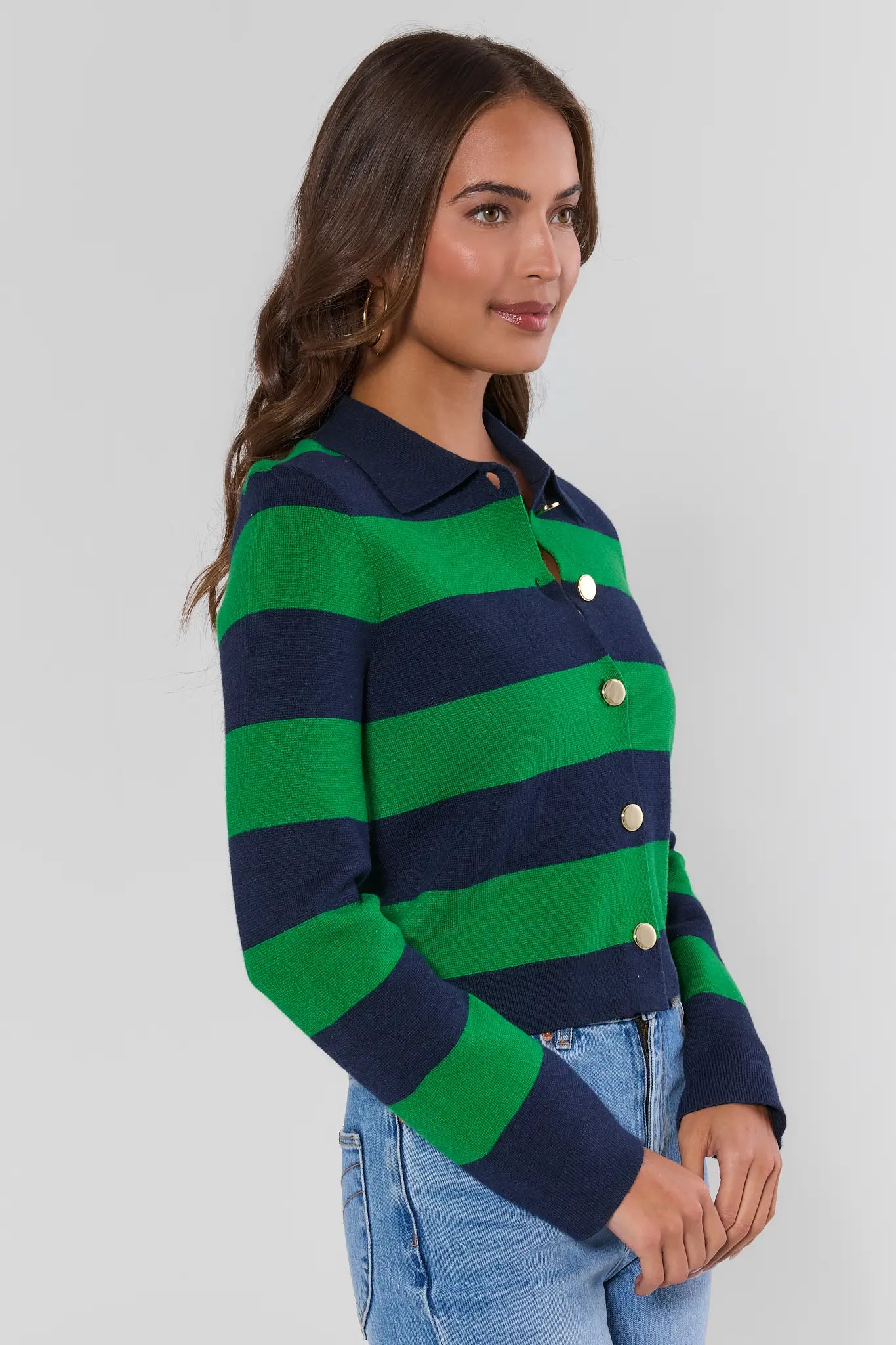 A woman wearing a green and navy blue striped knit cardigan with gold buttons, paired with light wash blue jeans, photographed against a neutral light gray studio background.