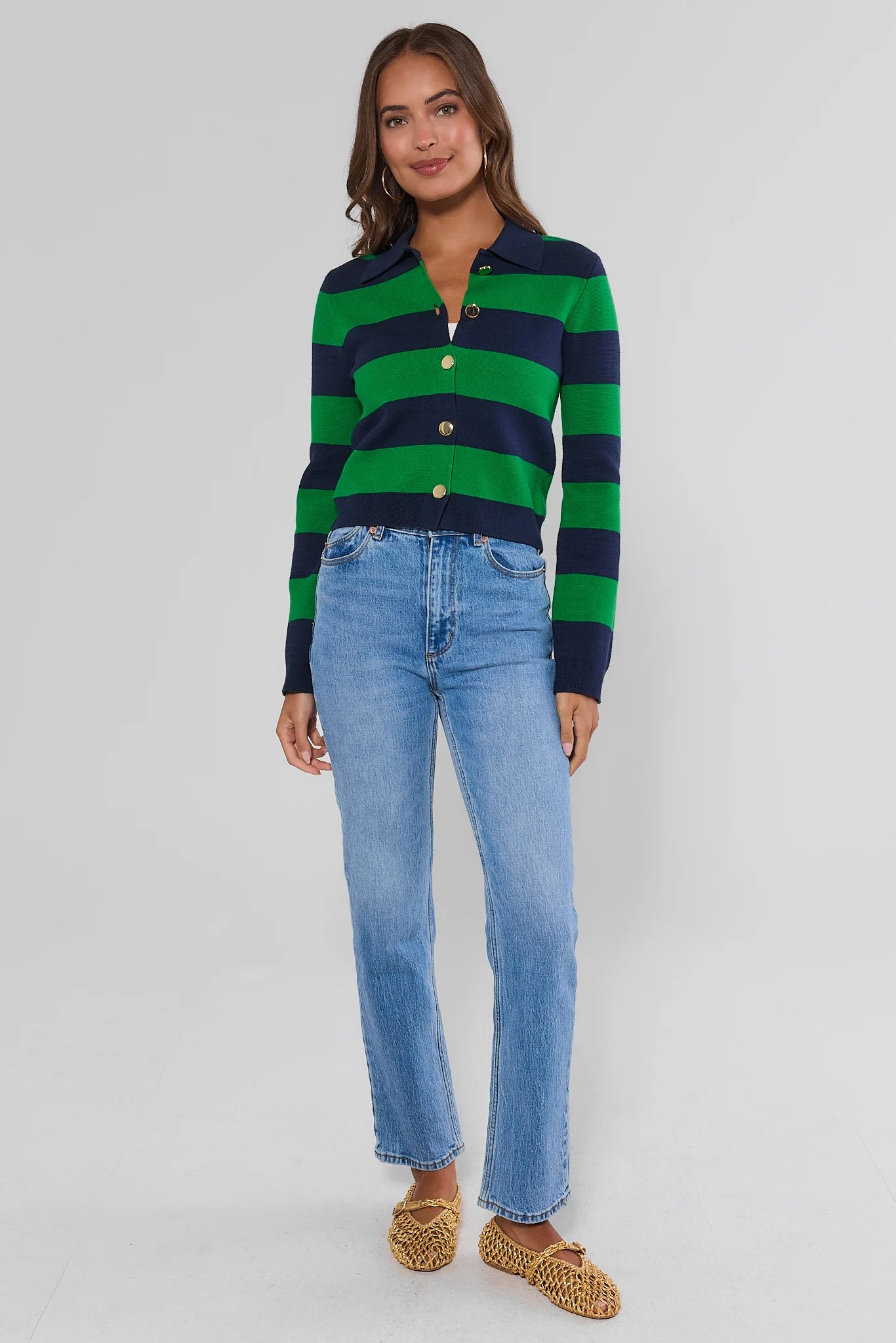 A woman modeling a green and navy blue horizontally striped knit polo cardigan with gold buttons, paired with light-wash blue jeans and brown woven flat shoes, against a light gray studio background.
