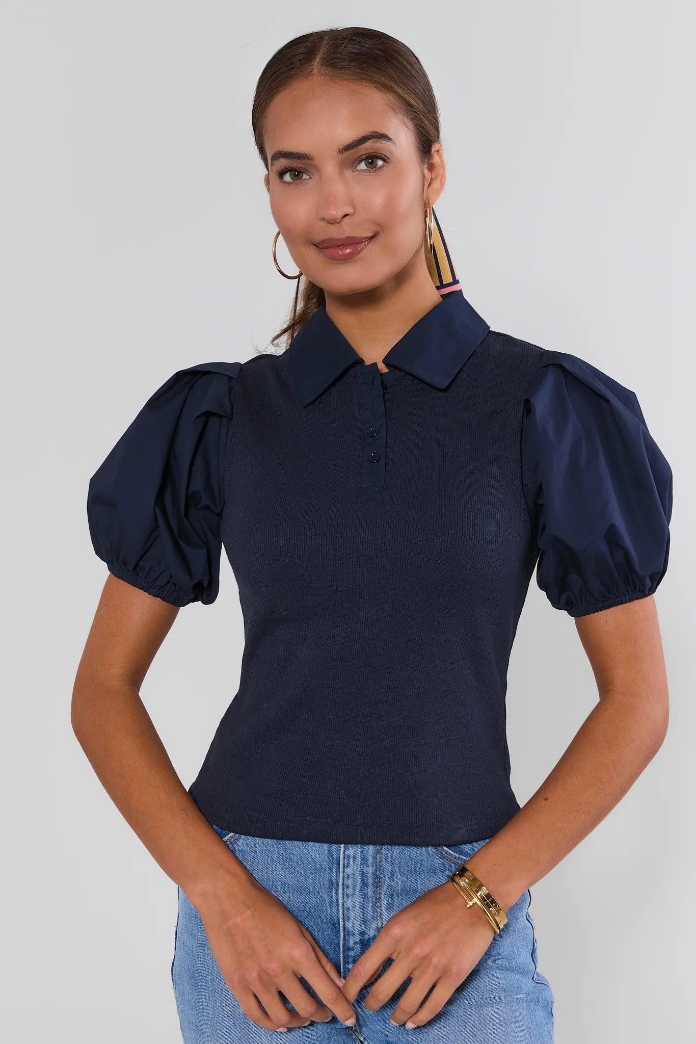 A woman wearing a navy blue short-sleeve polo shirt with voluminous puff sleeves and light blue jeans, photographed against a plain light gray studio background.