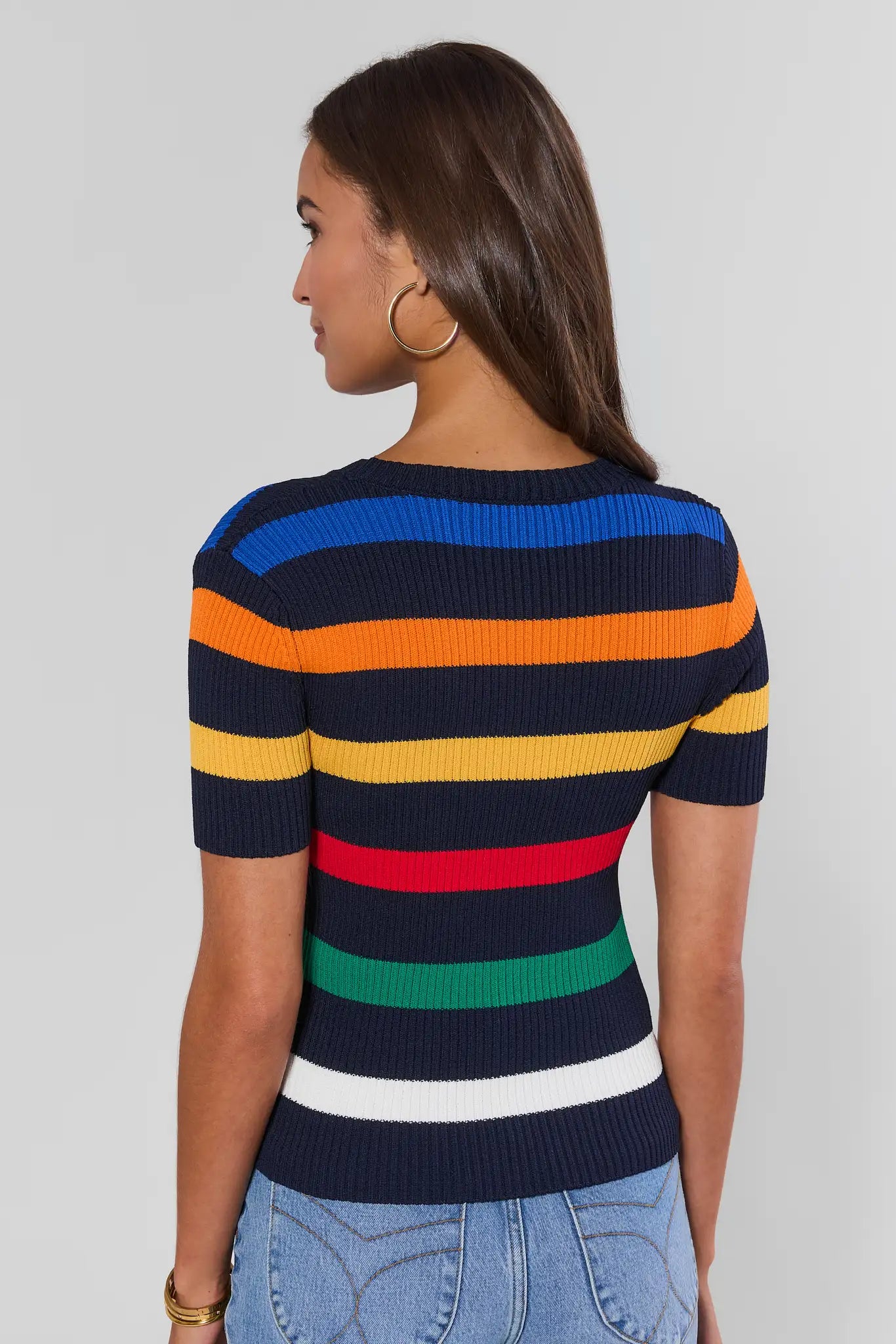 A woman shown from the back wearing a short-sleeve ribbed knit sweater with horizontal stripes in blue, orange, yellow, red, green, white, and navy, paired with light blue jeans against a light gray studio background.