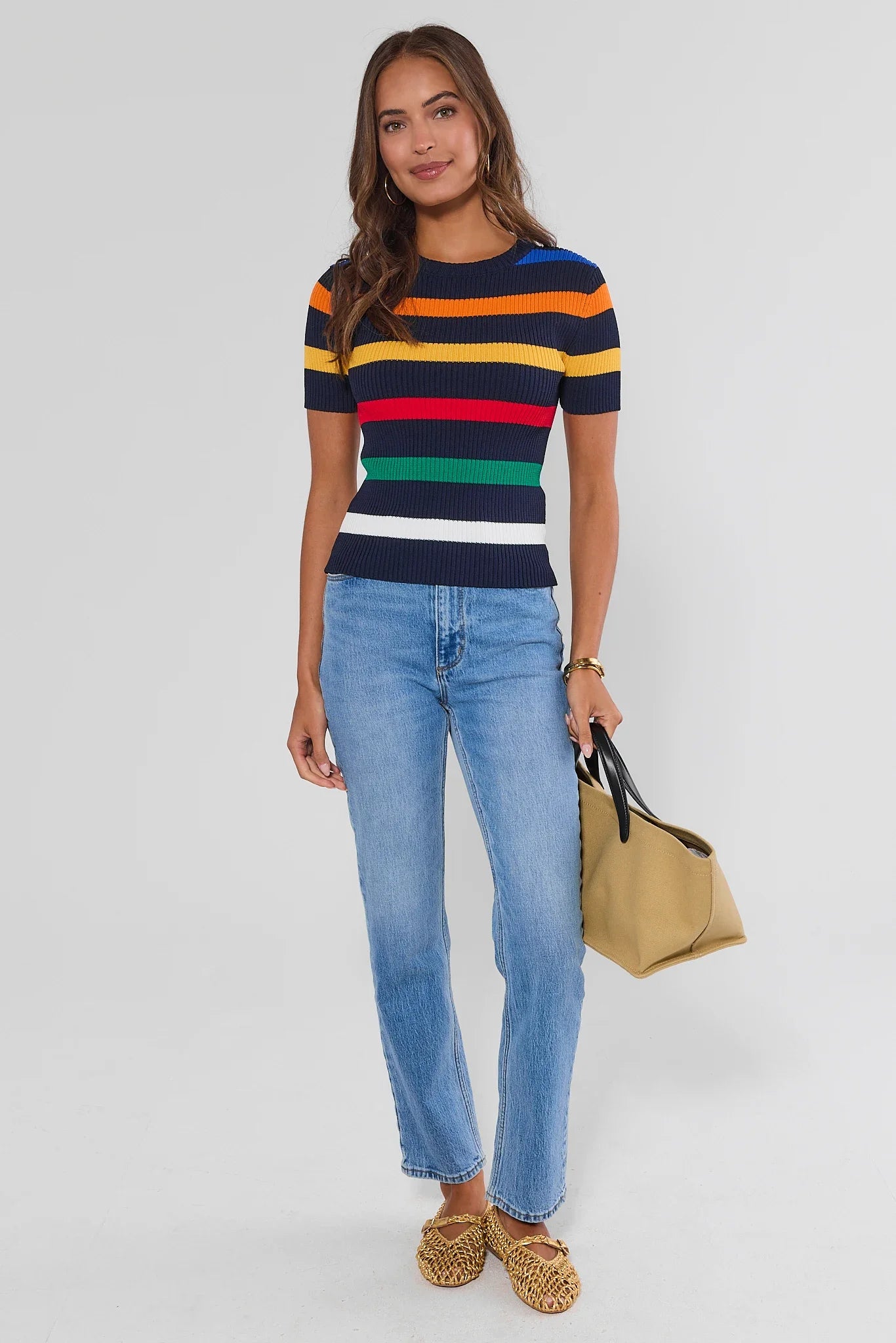 A woman modeling a short-sleeve knit top with horizontal navy, yellow, orange, red, green, and white stripes, paired with light-wash blue jeans and holding a tan tote bag against a light gray studio background.