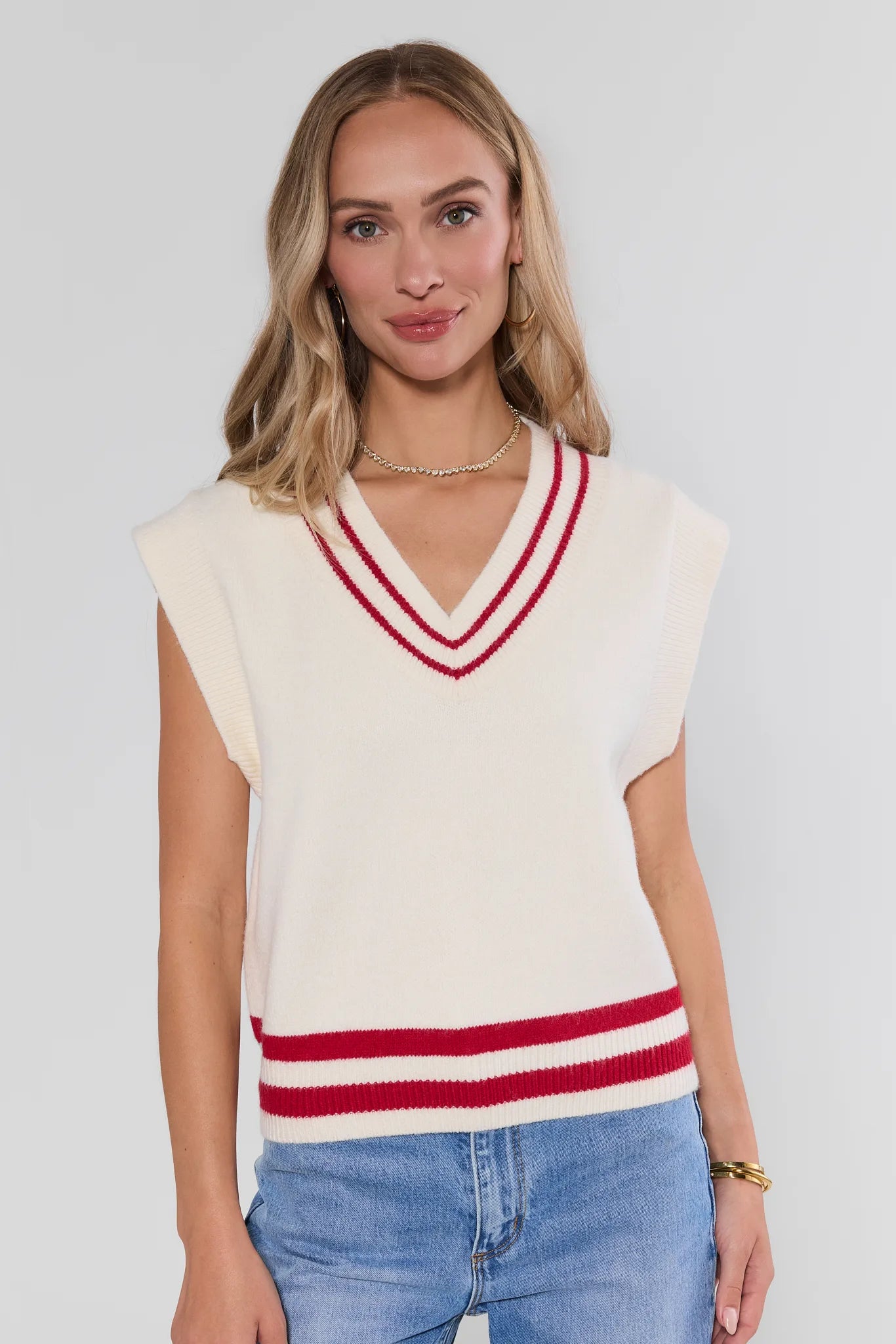A woman wearing a cream-colored sleeveless knit sweater vest with red double stripes on the V-neck and hem, paired with blue jeans, against a light gray studio background.