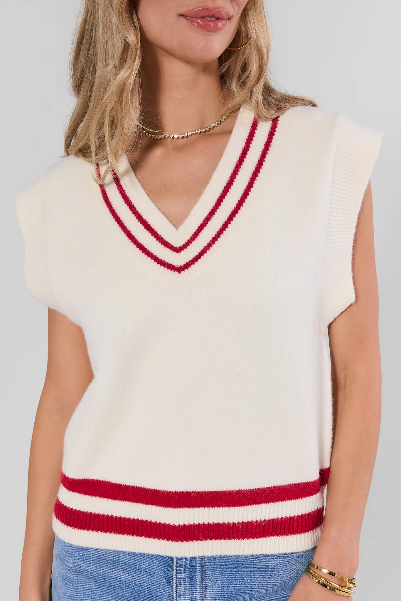 A woman wearing a cream knit sleeveless sweater vest with red double stripes on the V-neck and hem, paired with blue jeans against a light gray background.