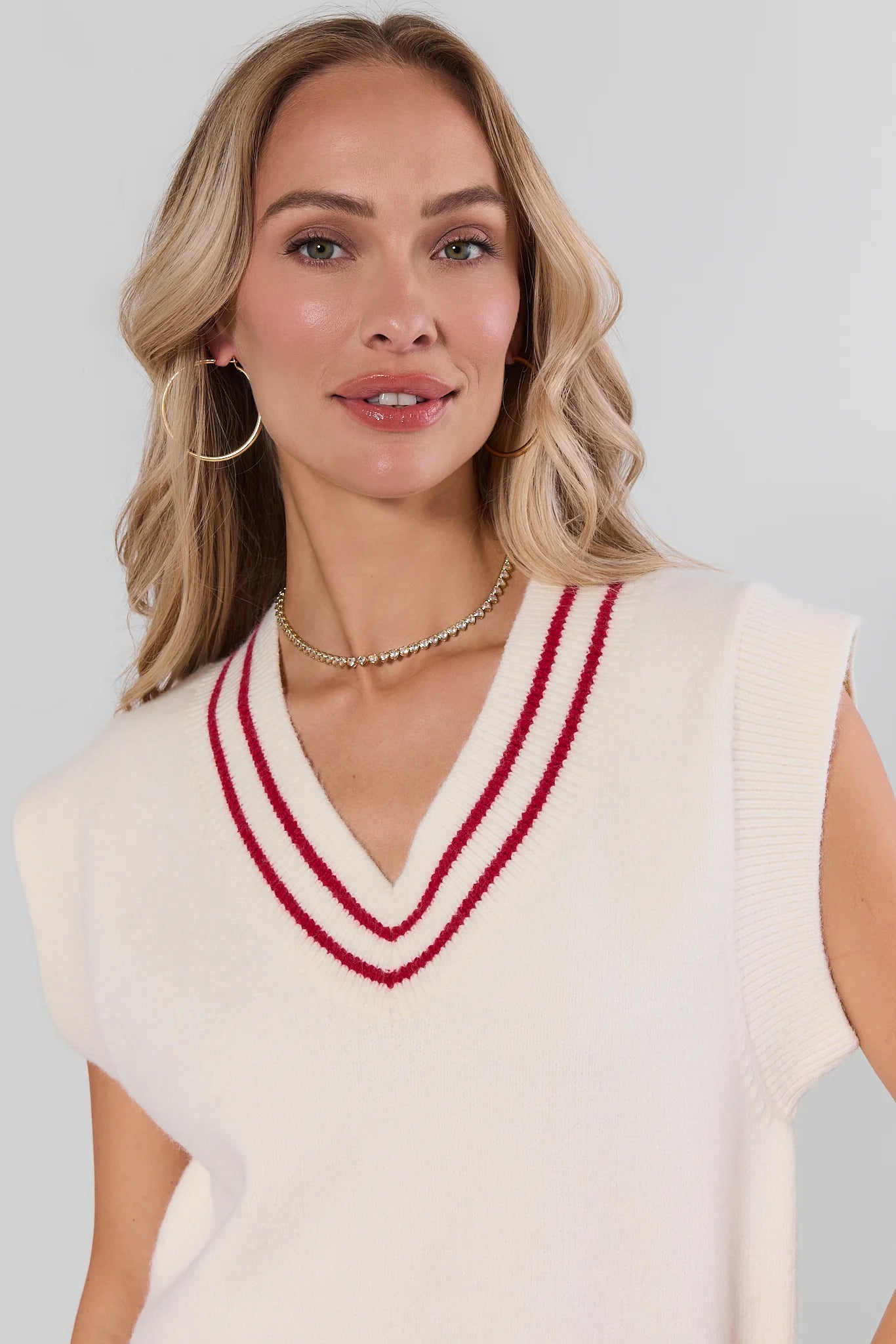 A woman wearing a cream-colored sleeveless knit sweater vest with a red and navy striped V-neck trim, paired with gold hoop earrings and a beaded necklace, against a light gray studio background.