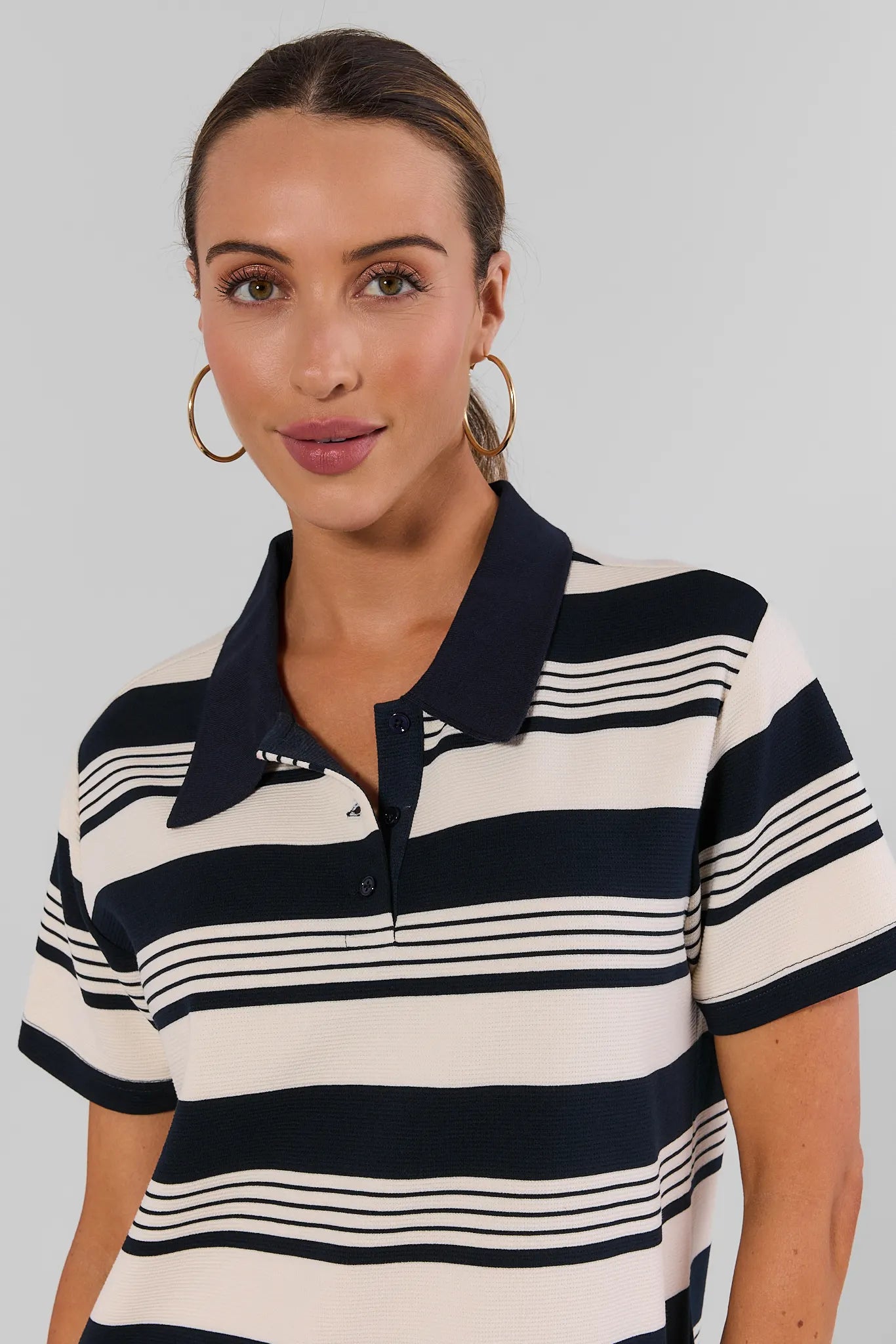 A woman wearing a short-sleeve polo shirt with horizontal navy and cream stripes, paired with gold hoop earrings, against a solid light gray background.