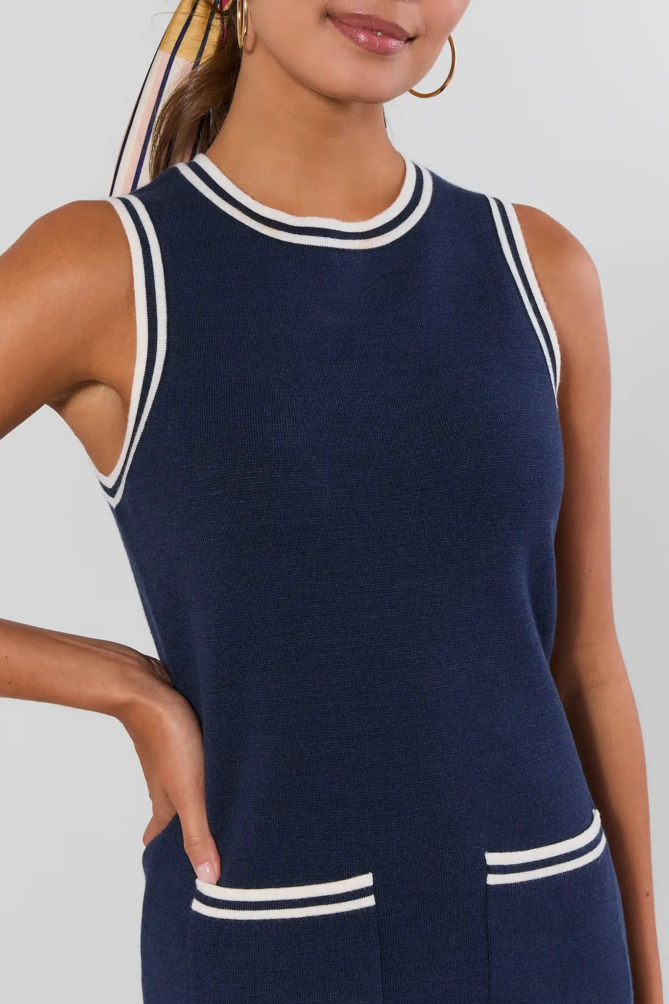 A close-up of a woman wearing a navy sleeveless knit top with white double-trimmed collar and pocket bands, against a light gray studio background.