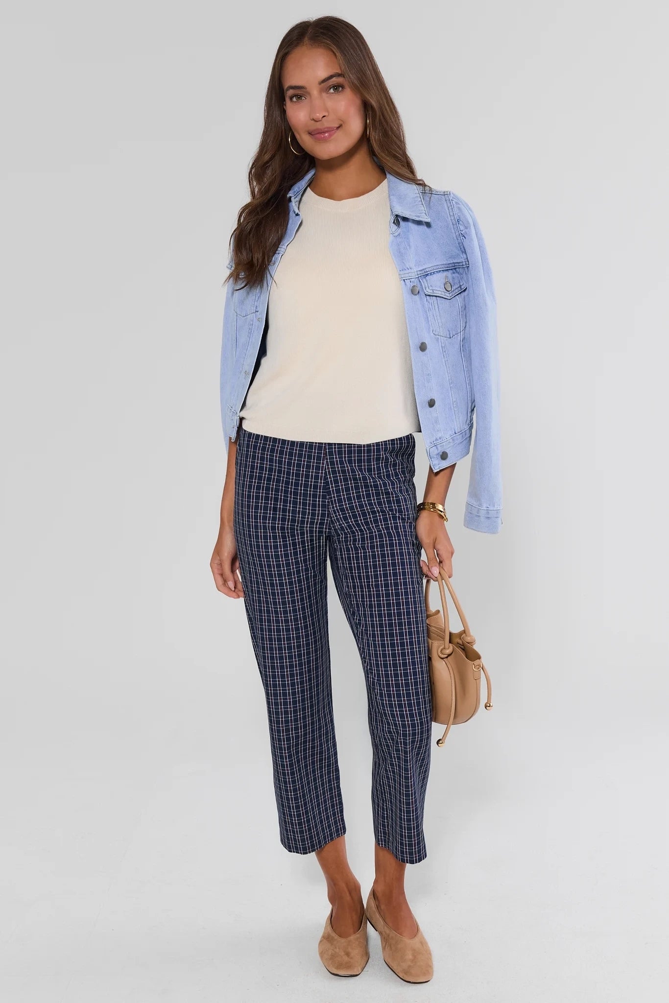 A woman modeling a light blue denim jacket over a cream ribbed top, paired with navy and black checkered cropped trousers, beige suede shoes, and holding a small tan handbag against a light gray studio background.