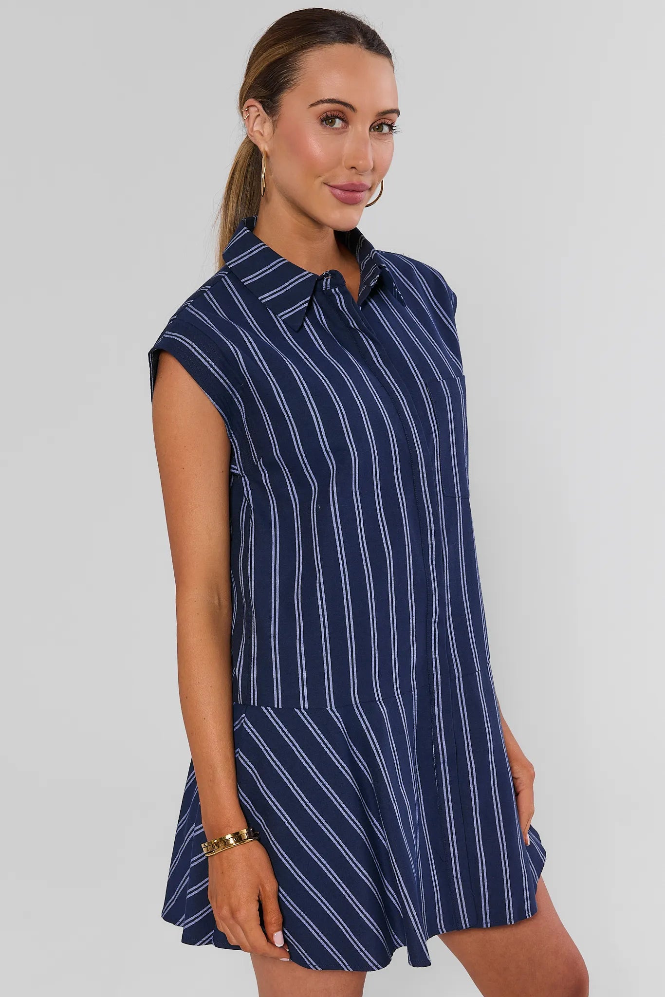 A woman modeling a sleeveless navy blue shirt dress with thin white vertical stripes, featuring a pointed collar and a flared skirt, set against a plain light gray background.