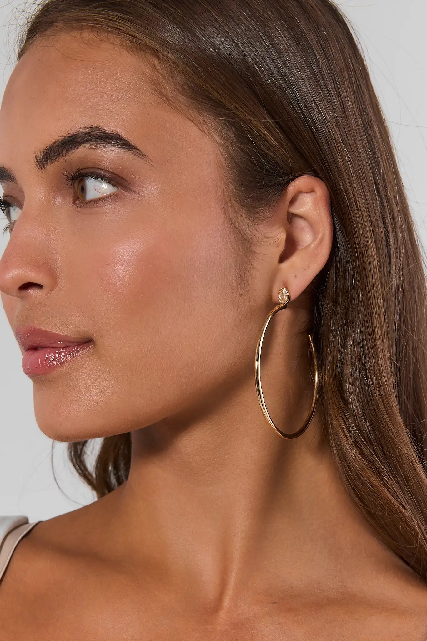 A close-up side profile of a woman wearing a pair of large gold hoop earrings with a small gold stud earring, against a light gray background.