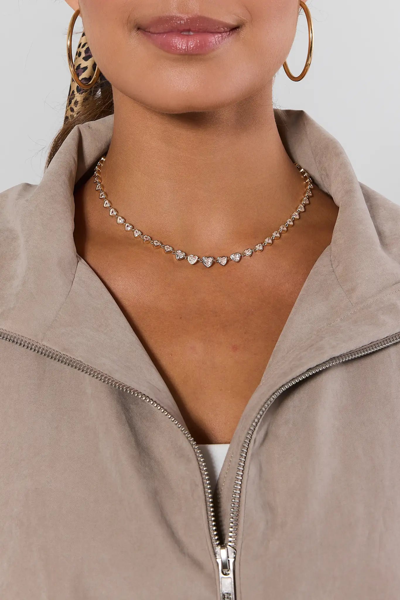 A close-up of a model wearing a silver-toned heart-link choker necklace, styled with a beige zip-up jacket and gold hoop earrings.