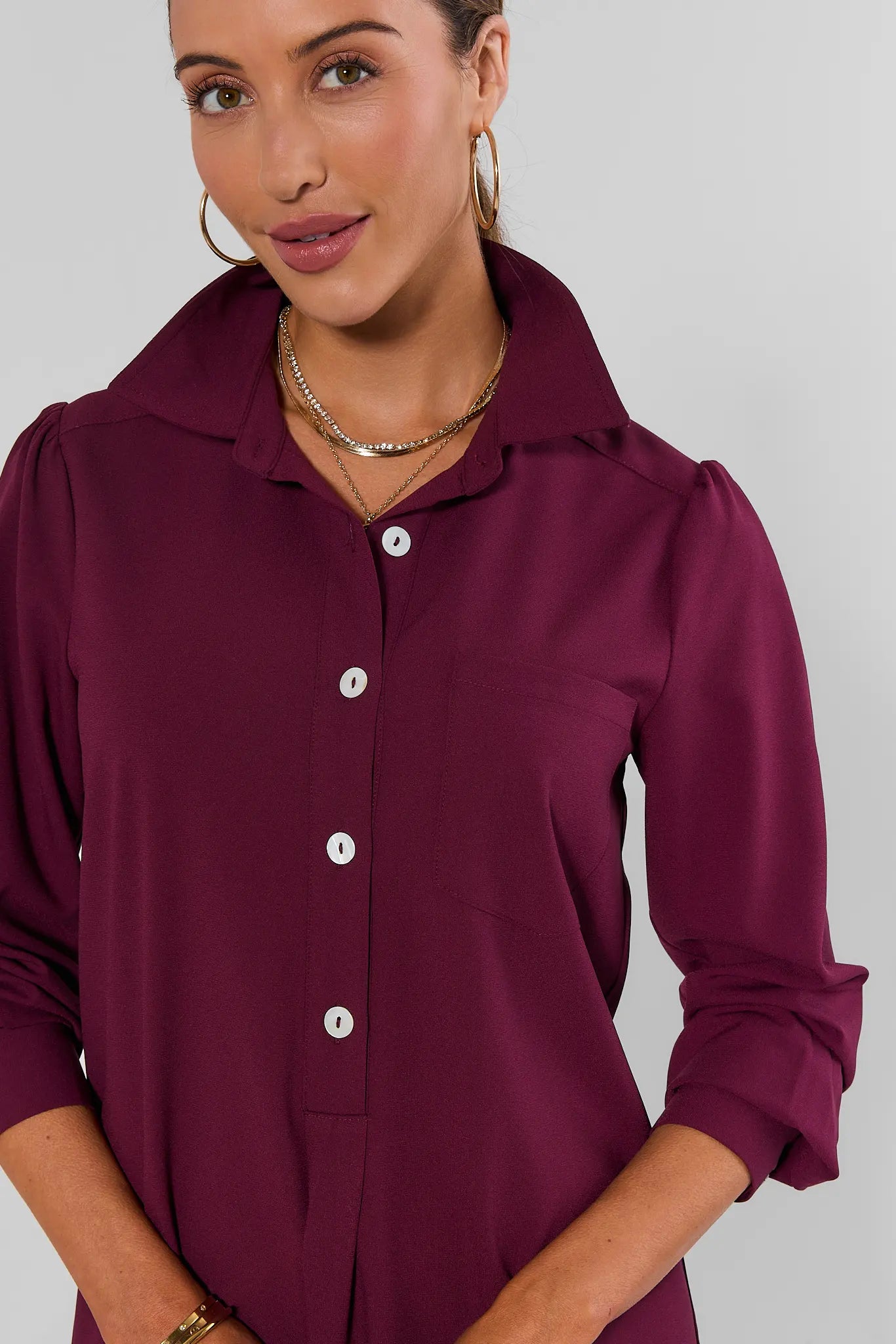 A woman wearing a long-sleeve button-up shirt in deep burgundy with white buttons, shown from the chest up against a plain light gray studio background.