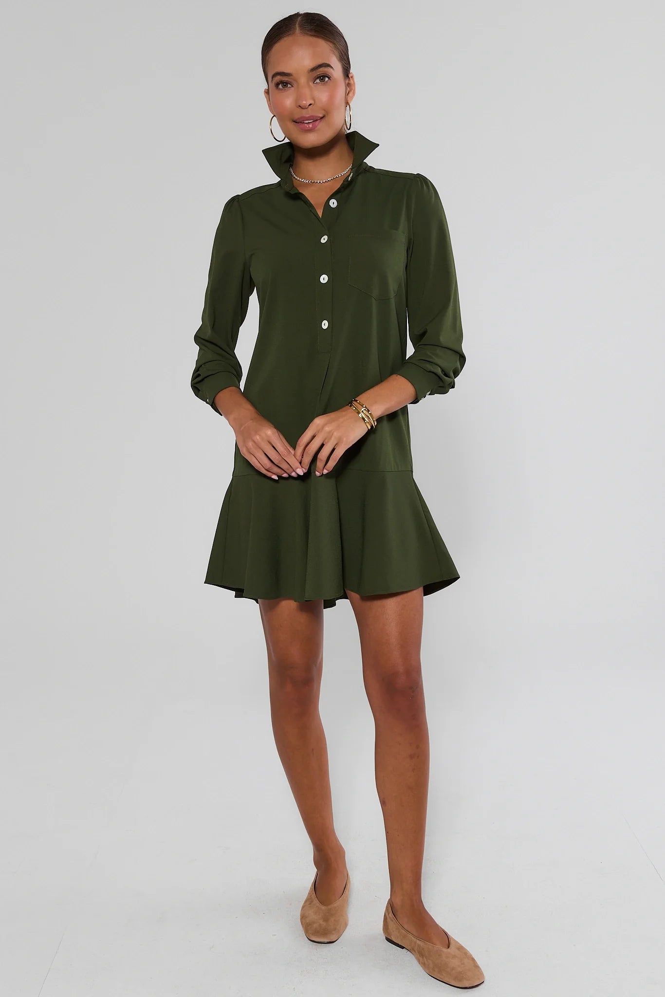 A woman modeling an olive green long-sleeve mini dress with a button-down front and a flared hem, paired with tan flats, against a light gray background.