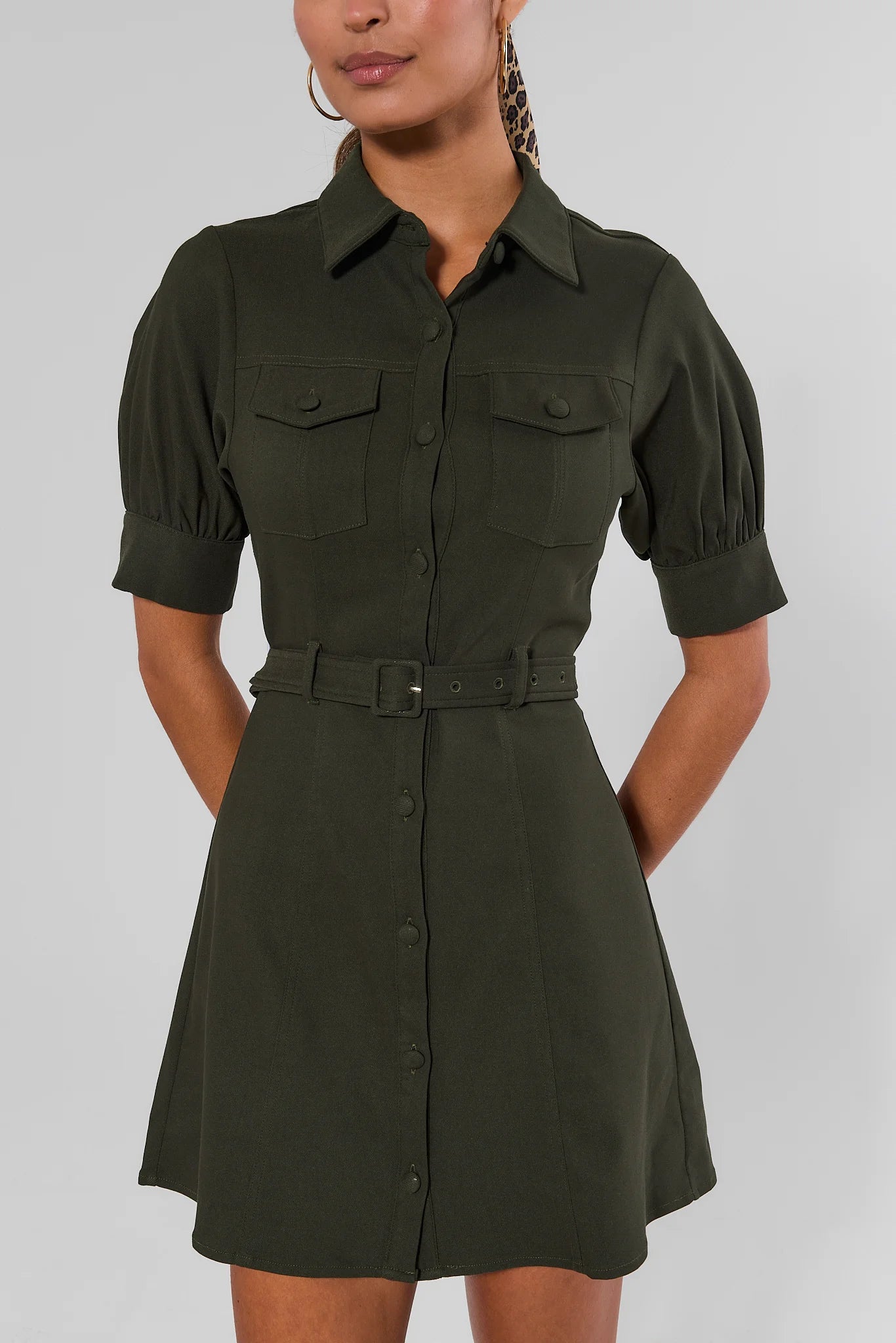 A woman wearing an olive green short-sleeve shirt dress with puff sleeves, a button-down front, chest flap pockets, and a matching belt, photographed against a light gray studio background.