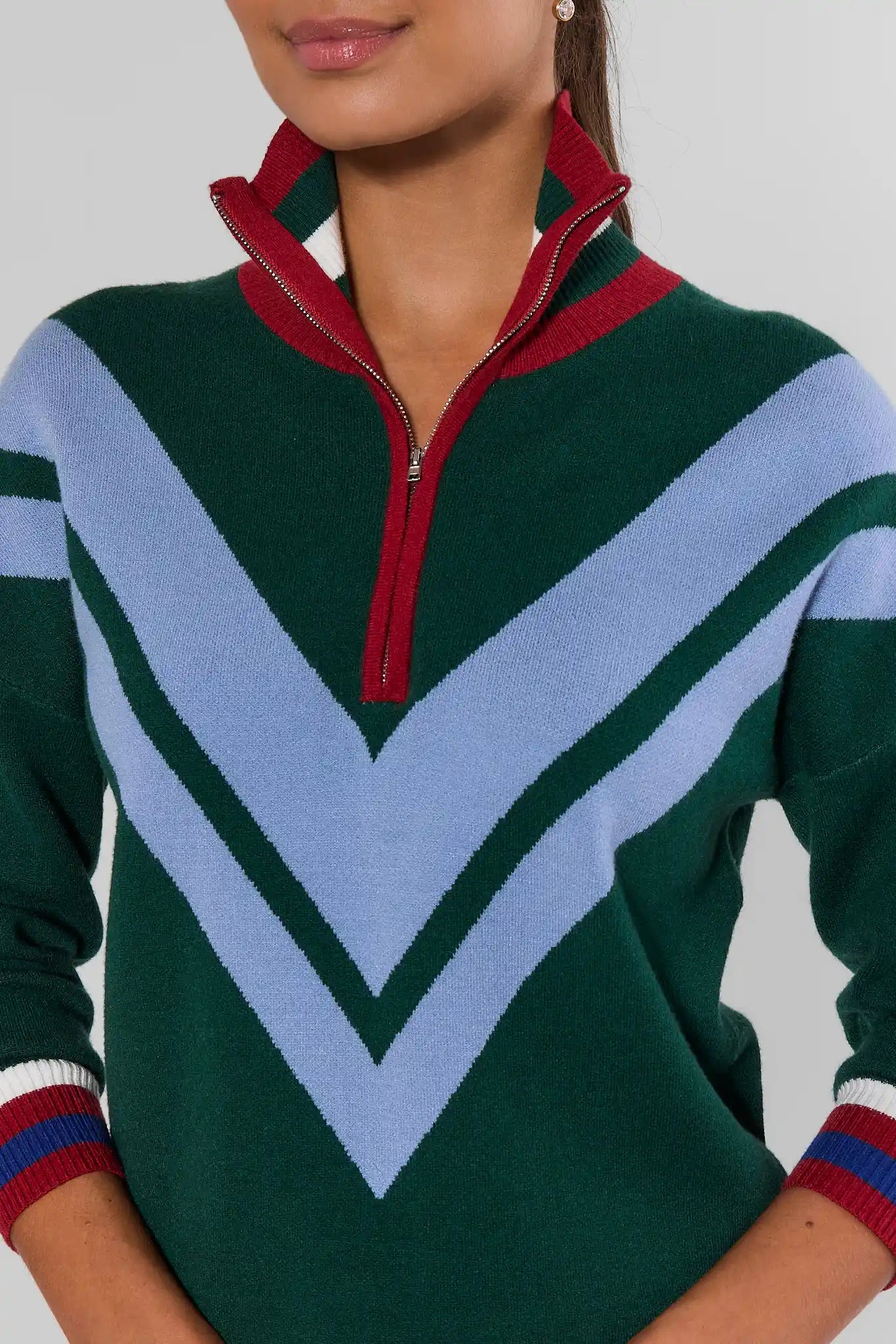 A close-up of a woman wearing a green quarter-zip sweater with a red collar and zipper trim, featuring light blue chevron stripes across the front.