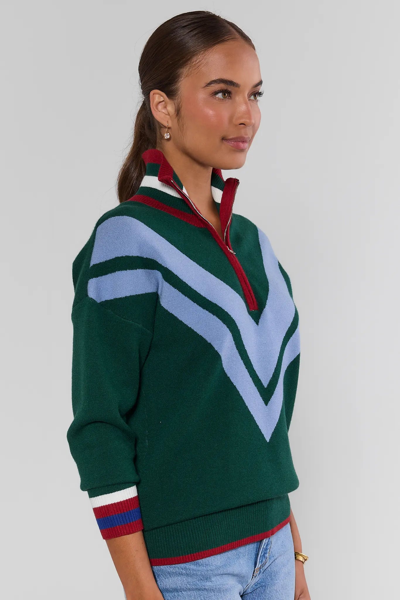 A woman wearing a green knit half-zip sweater with light blue and white chevron patterns and red and white striped collar and cuffs, paired with light blue jeans, against a plain light gray background.