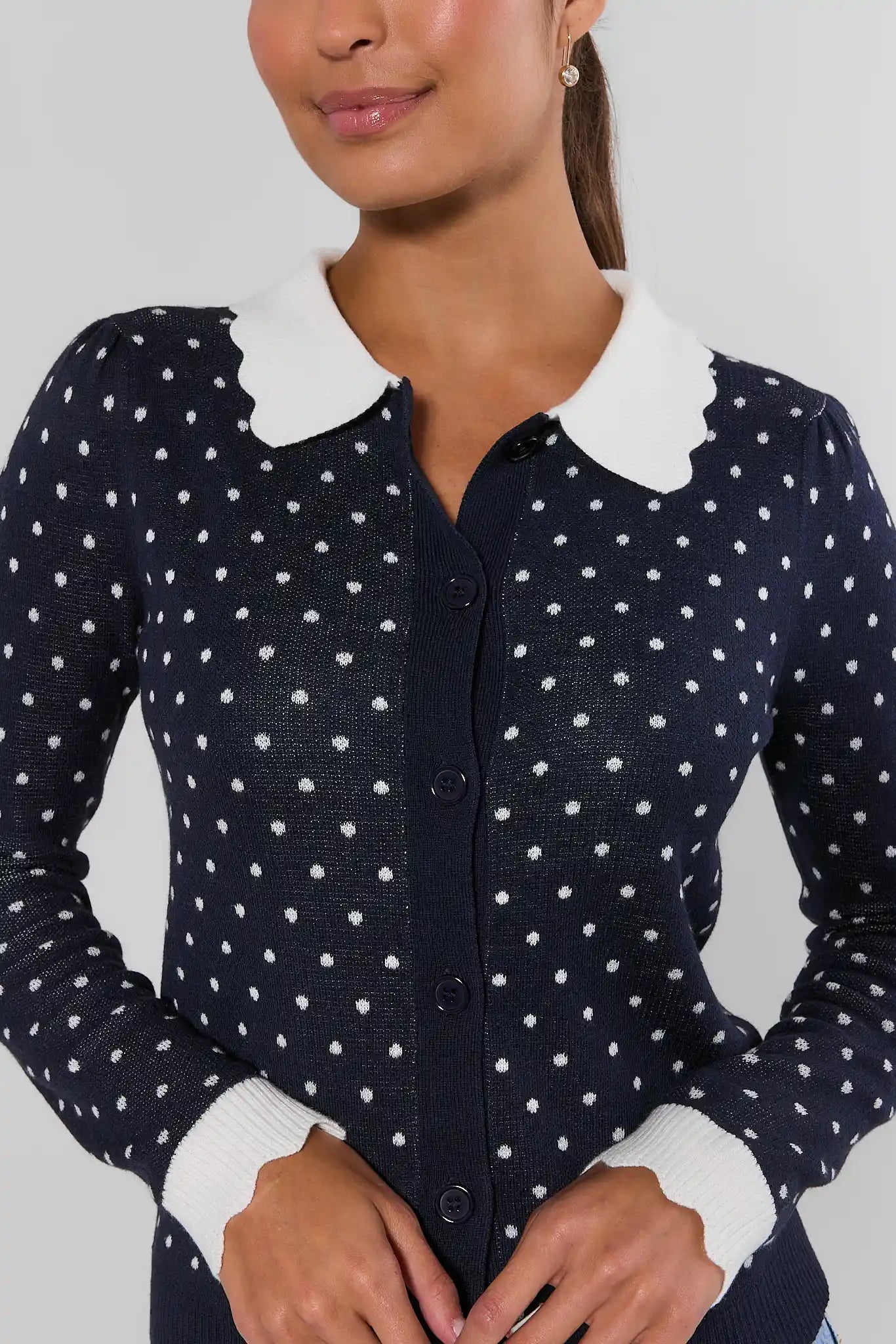 A woman wearing a navy blue long-sleeve cardigan with white polka dots and a contrasting white Peter Pan collar, shown from the chest up against a light gray background.