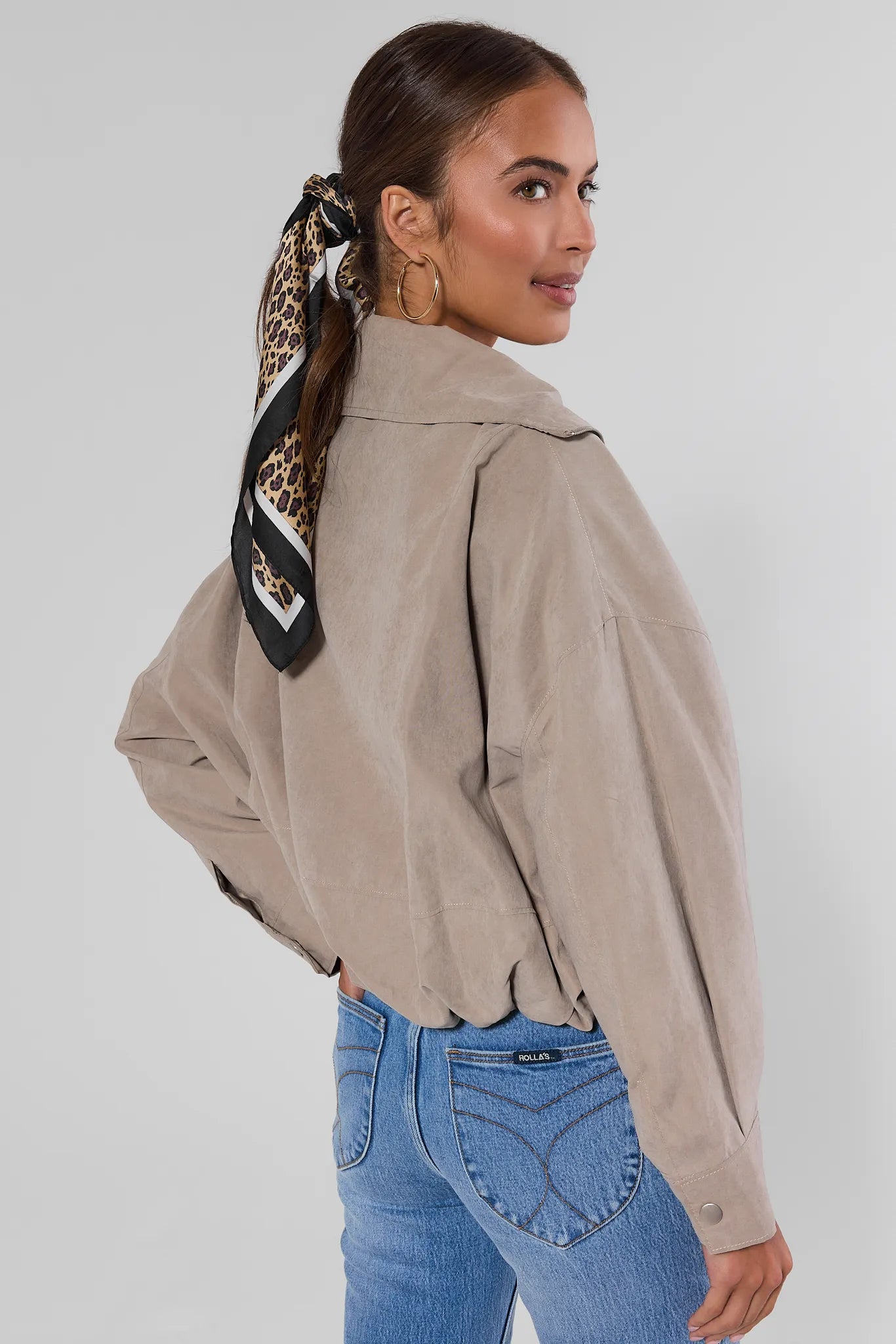 A woman wearing a light taupe-brown cropped utility jacket with a leopard-print neck scarf, paired with blue jeans, photographed from a three-quarters back view against a light gray studio background.