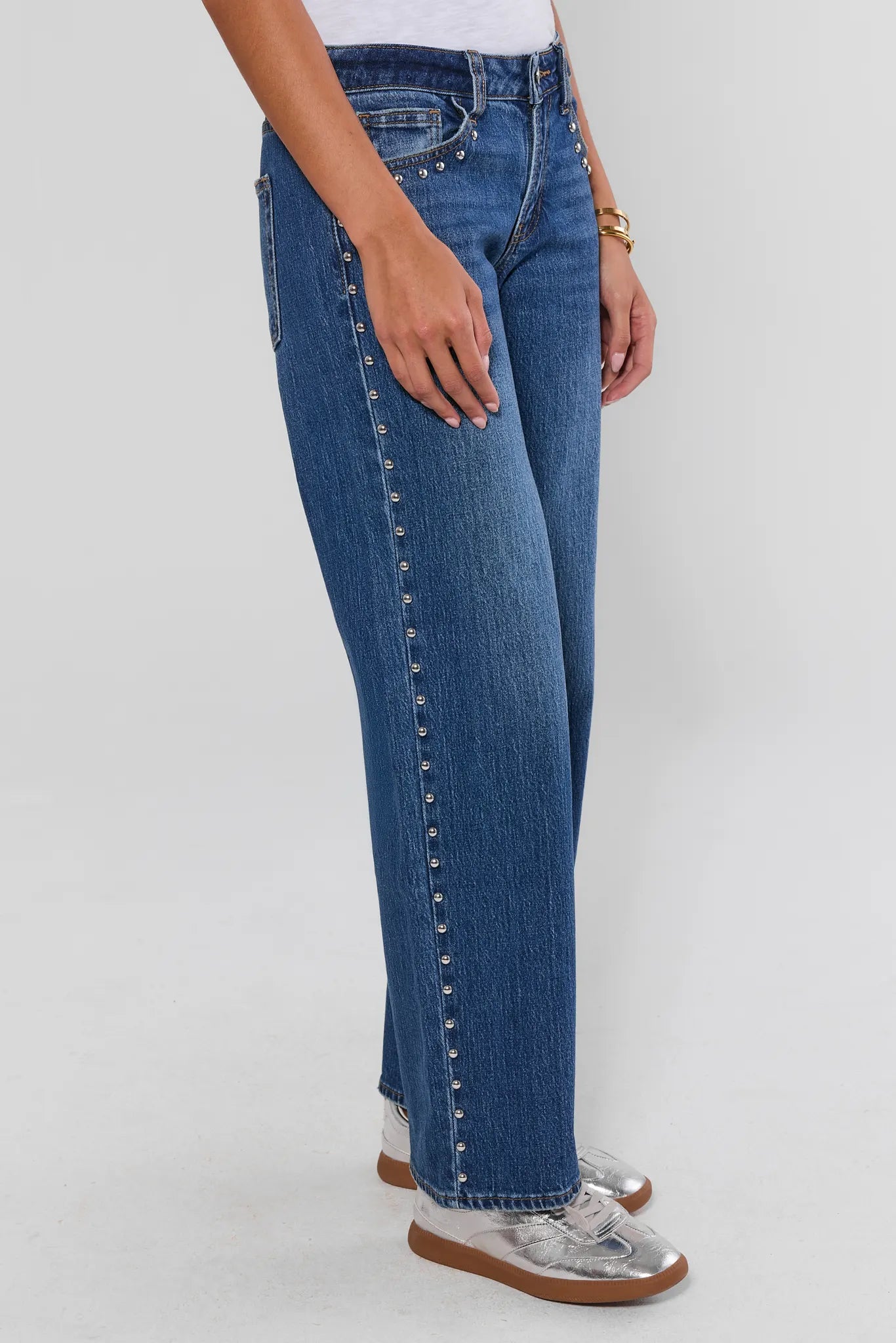 A person wearing blue denim jeans with silver stud detailing running down the outer side of the legs, paired with clear slip-on shoes, shown against a light gray studio background.