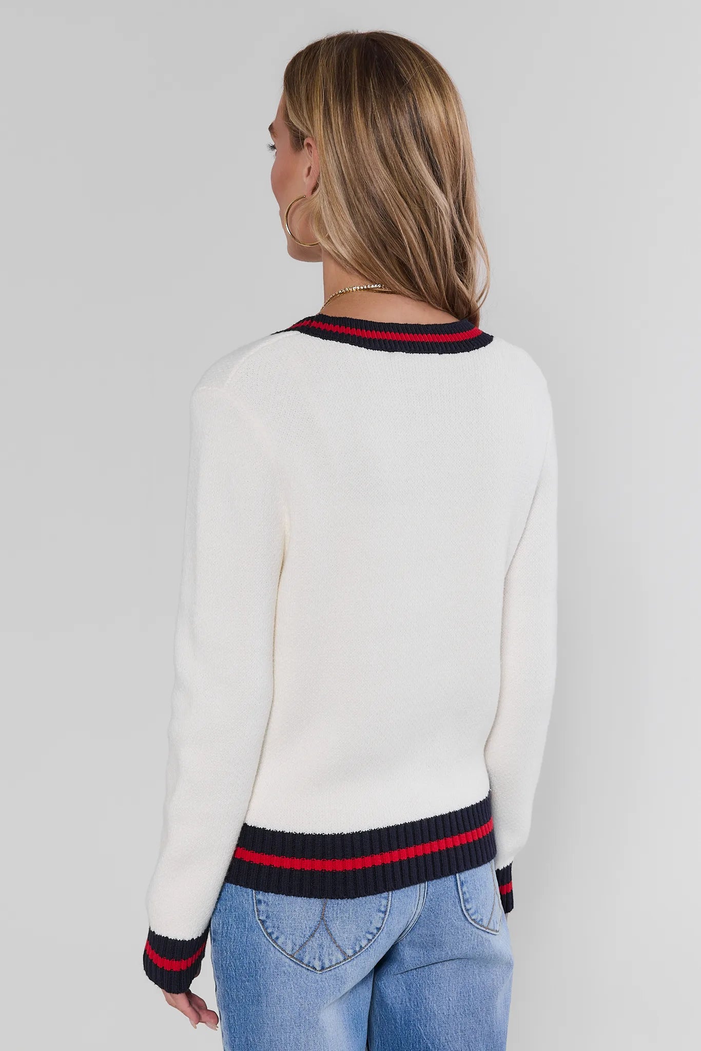 A woman shown from the back wearing a white knit sweater with navy and red striped ribbed trim at the crew neck, cuffs, and hem, paired with blue jeans against a light gray studio background.