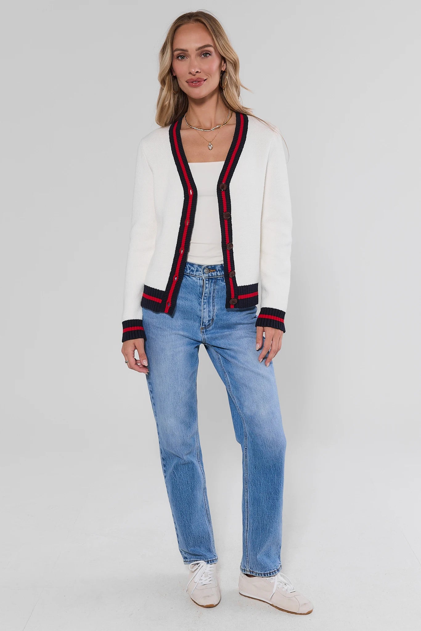A woman modeling a white knit cardigan with red and navy striped trim, paired with light blue jeans and white sneakers, against a light gray studio background.