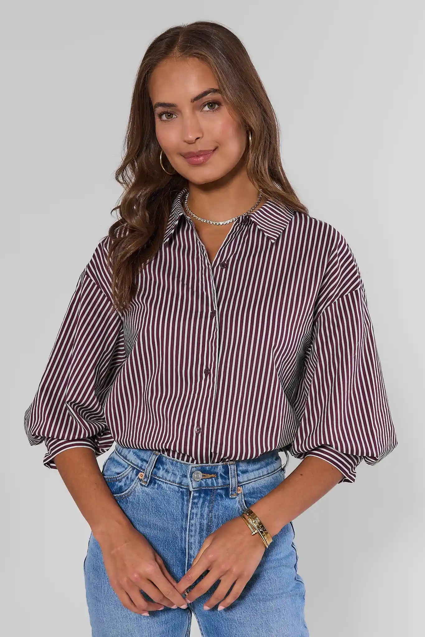A woman wearing a long-sleeve button-up shirt with vertical burgundy and white stripes, paired with light blue jeans, against a plain light gray background.
