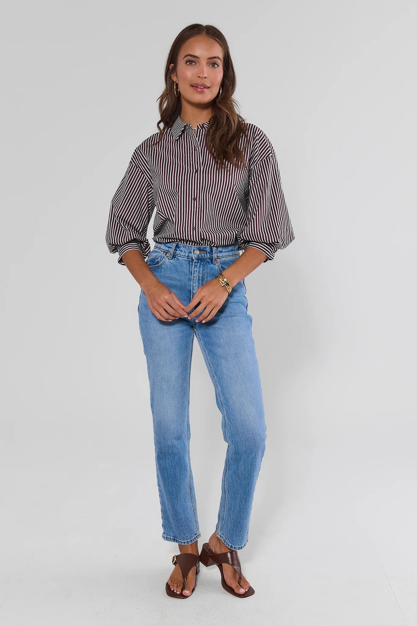 A woman modeling a long-sleeve button-up shirt with thin vertical stripes in dark brown and off-white, paired with light-wash blue jeans and brown leather sandals, against a plain light gray studio background.