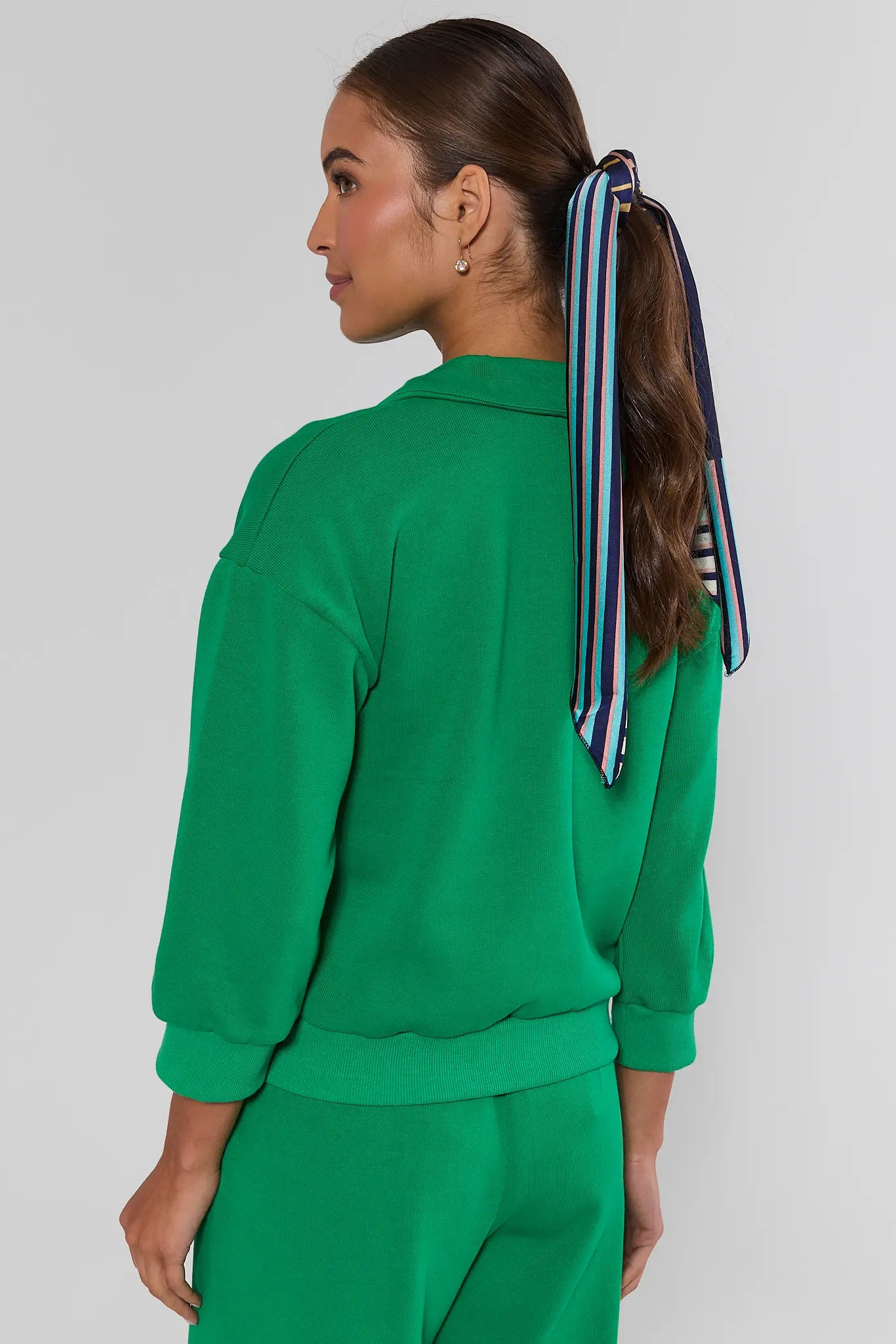 A woman shown from the back wearing a bright green sweatshirt and matching green pants, with a striped hair tie in her ponytail, against a light gray studio background.
